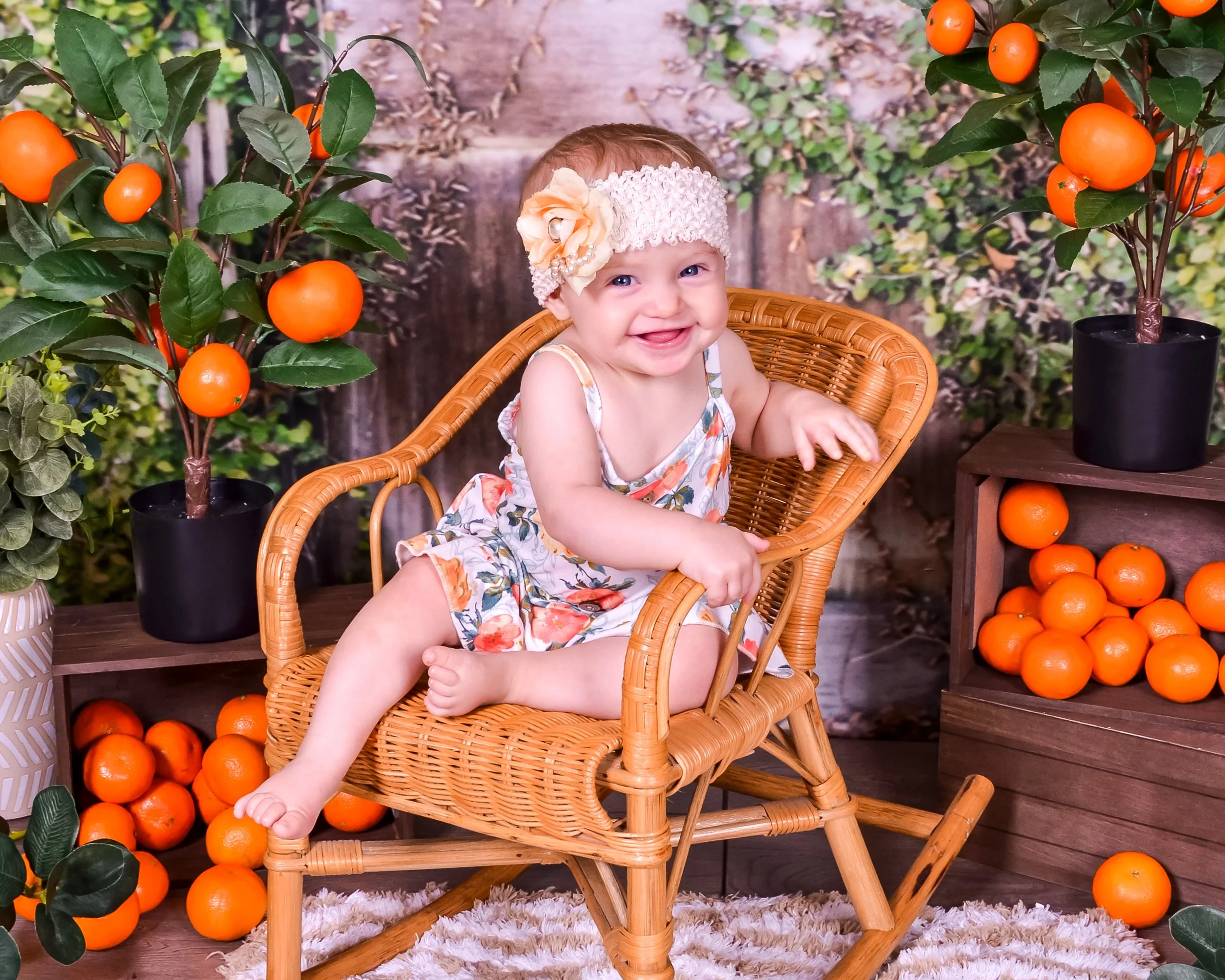A smiling baby girl sitting in a rattan rocking chair surrounded by potted orange trees and oranges.