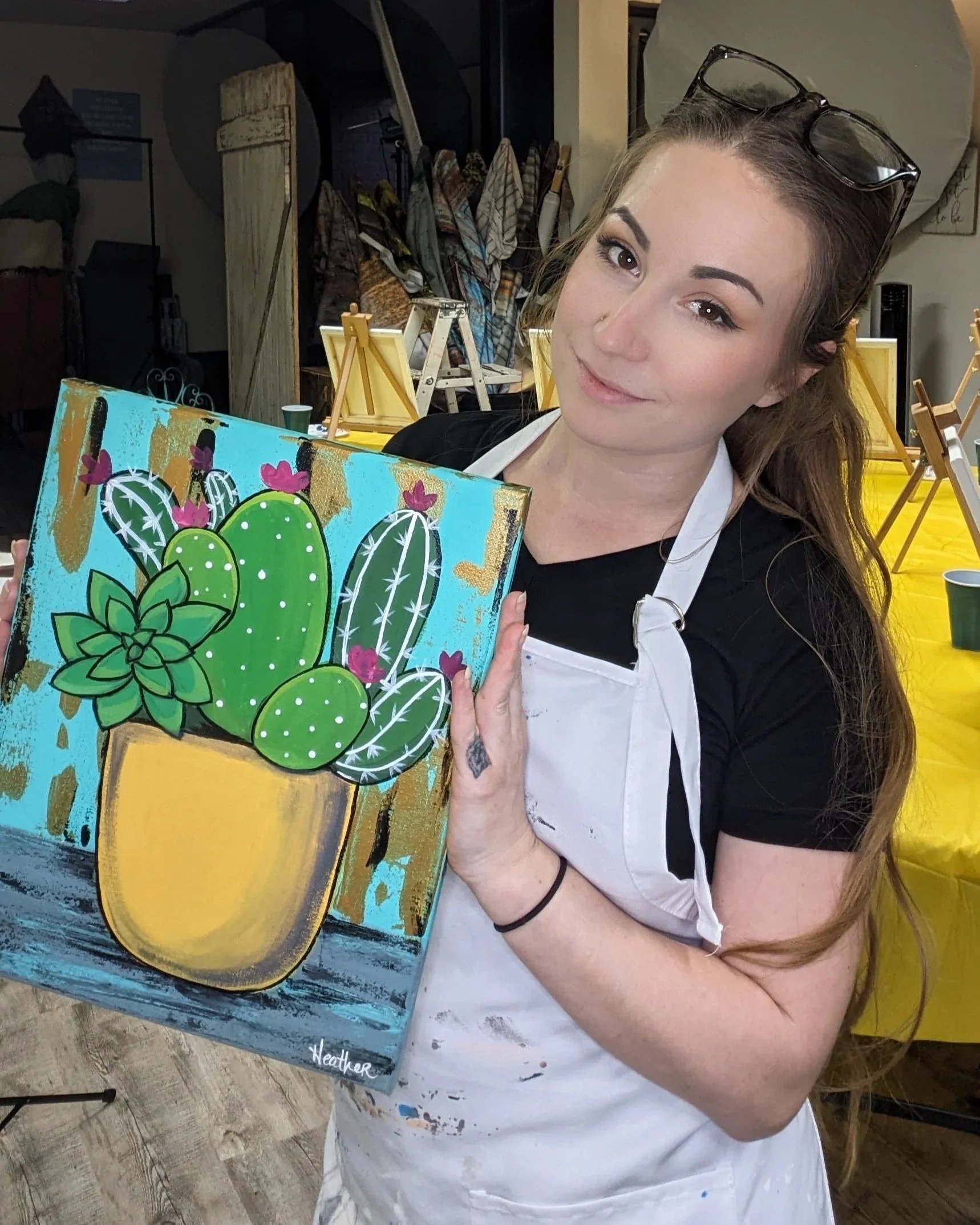 A woman with glasses on her head, holding a colorful painting of green cacti with pink flowers. She is wearing a white apron and has long brown hair. The background shows art supplies and easels in an art studio.