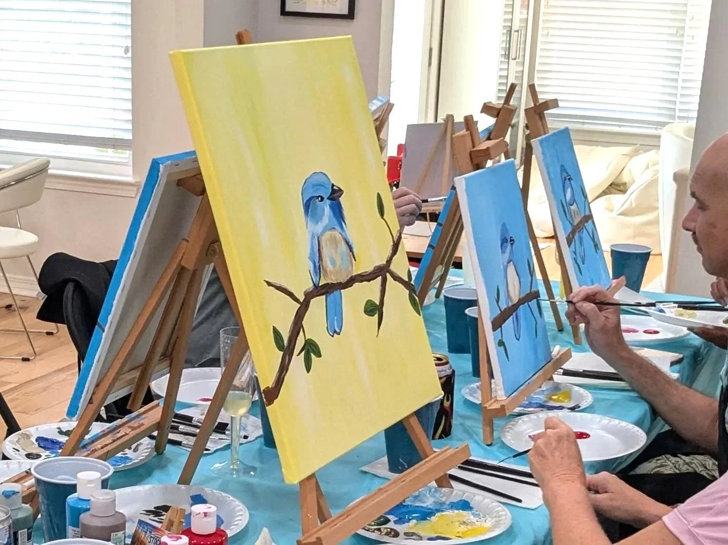 People painting blue birds on canvases at an indoor art class, with easels and painting supplies visible on the table.