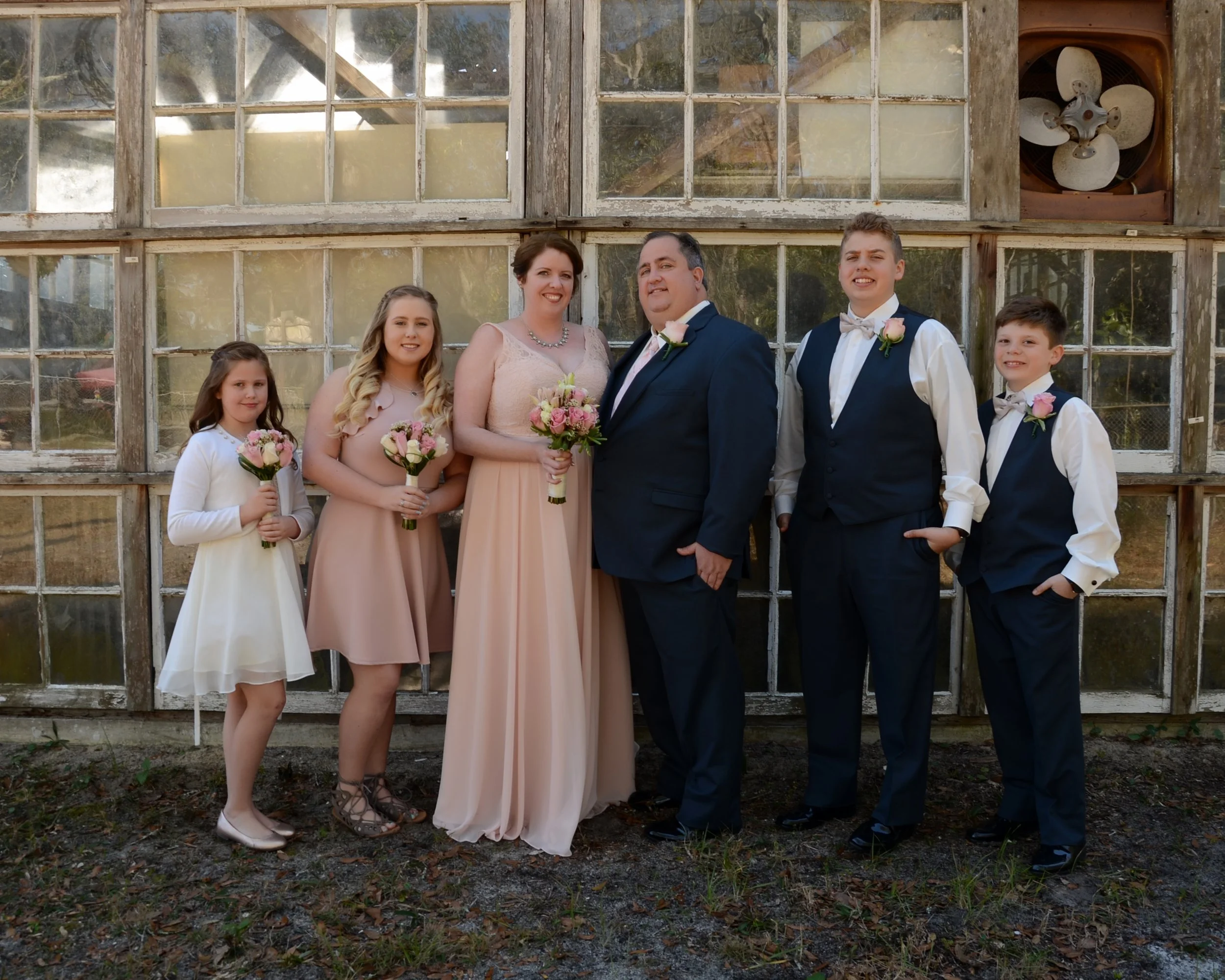 A wedding party of six standing outdoors in front of an old wooden building with large windows and a ventilator, all dressed in formal attire and holding bouquets of pink and white flowers.