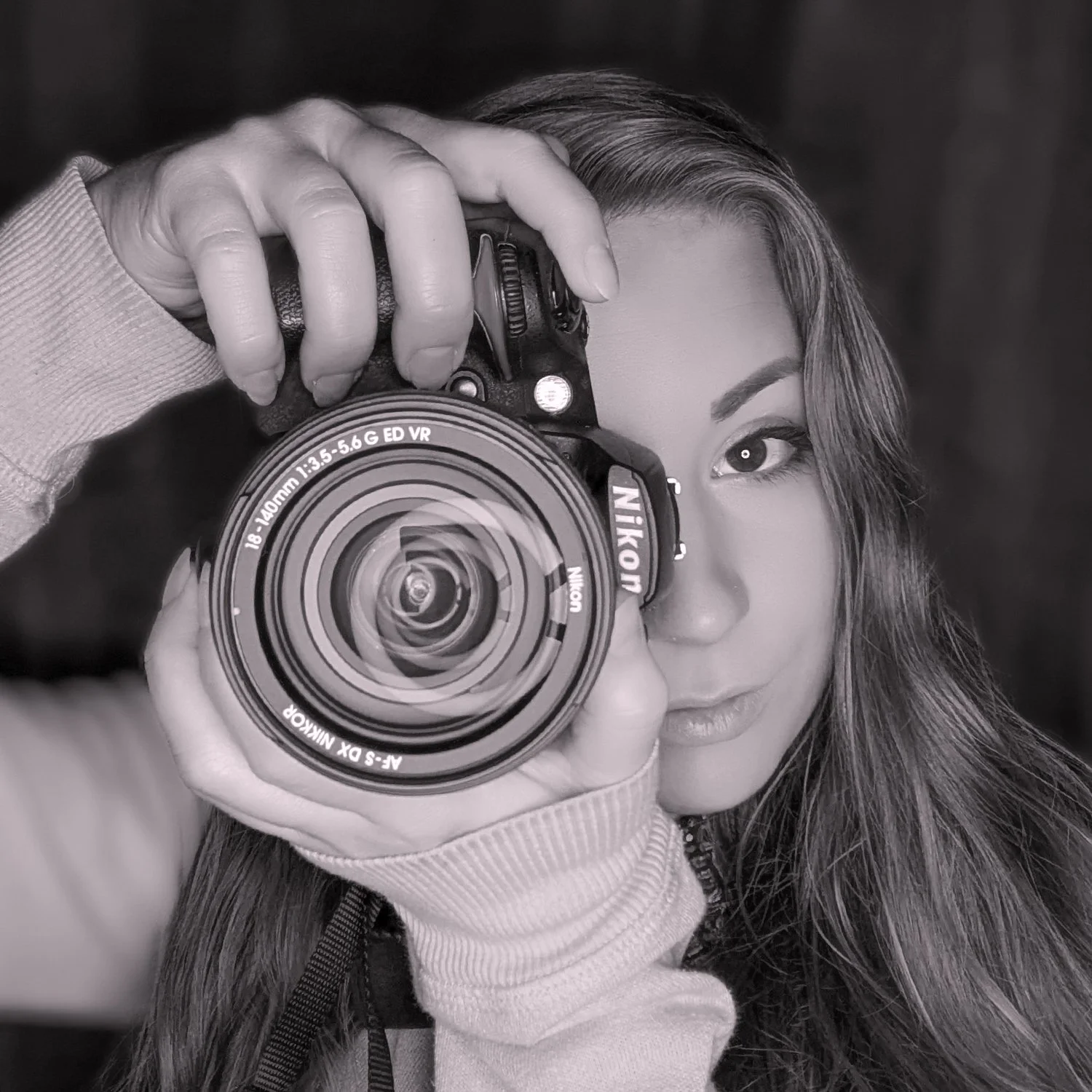A woman taking a selfie with a Nikon camera, partially covering her face, with her right eye visible and her long hair falling over her shoulder.