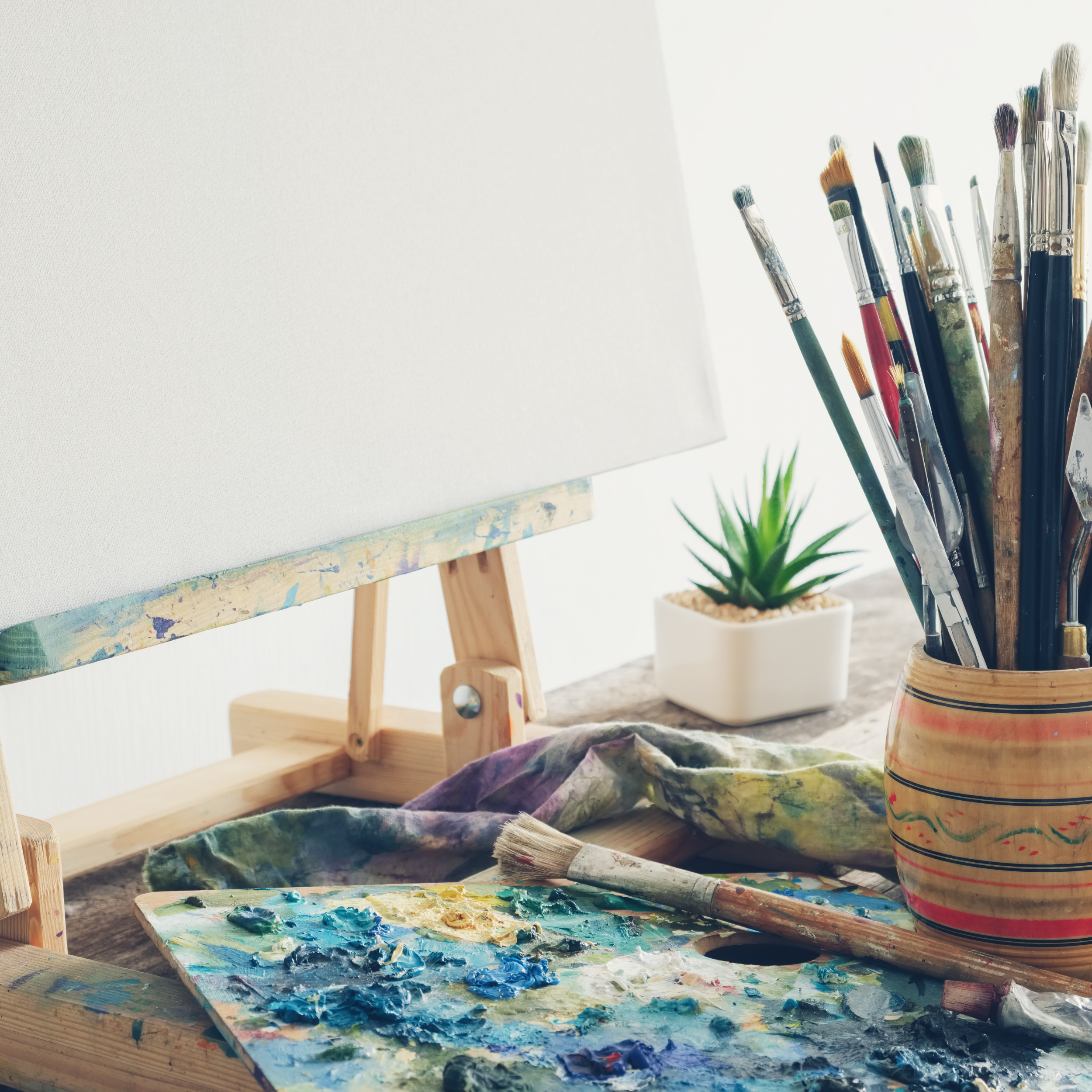 An artist's workspace with a blank canvas on a small wooden easel, a palette with blue and white paint, paintbrushes in a round wooden holder, a purple cloth with paint stains, and a small green aloe Vera plant in a white pot.