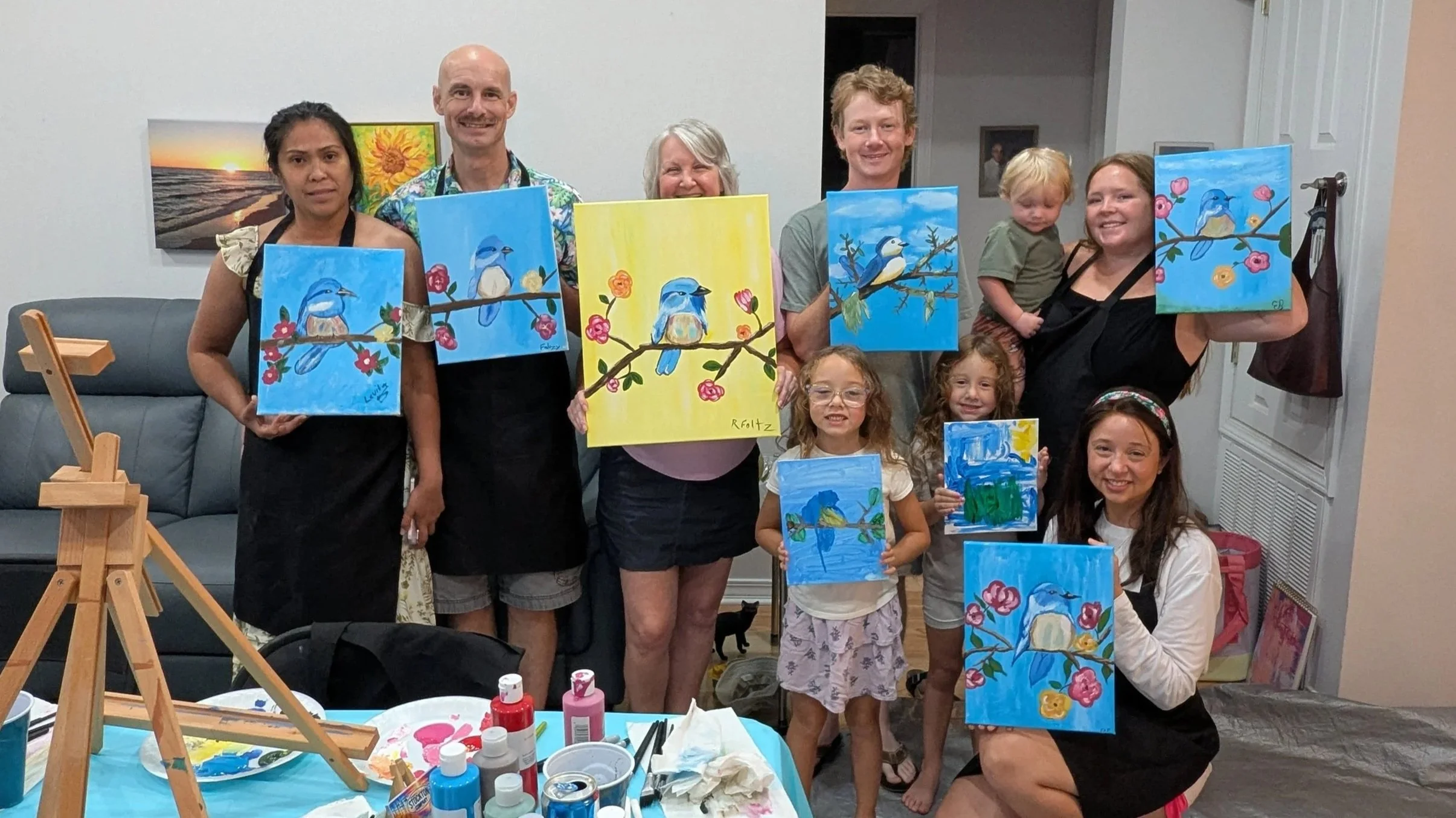 Group of people displaying blue and yellow bird-themed paintings at an art event in a room with white walls and artwork.