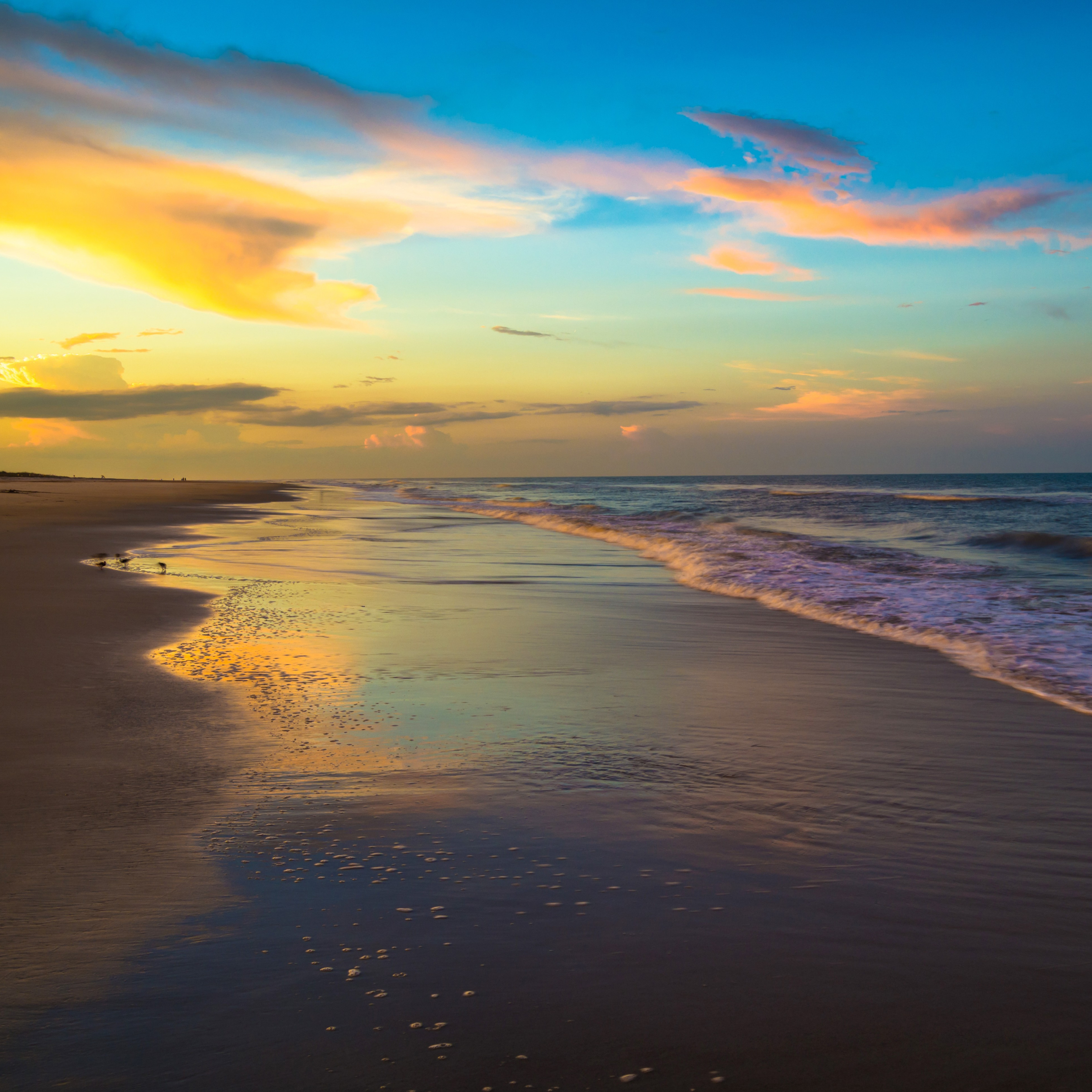 A scenic view of a beach during sunset, with colorful clouds in the sky, gentle waves hitting the shore, and the wet sand reflecting the sky's colors.