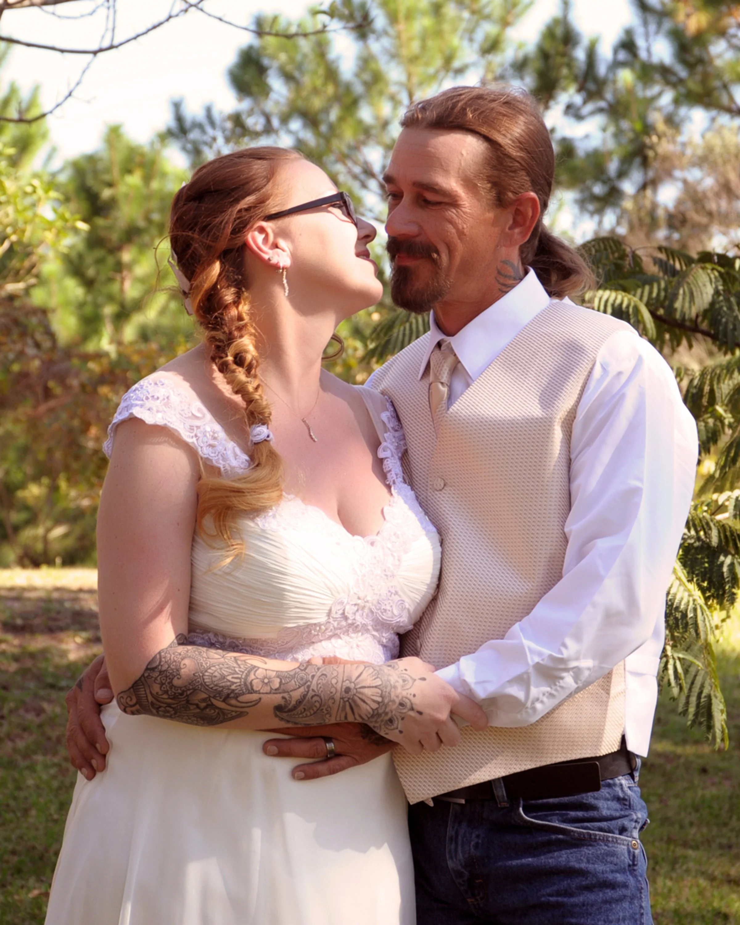 A bride and groom embrace outdoors on their wedding day, surrounded by trees and greenery.