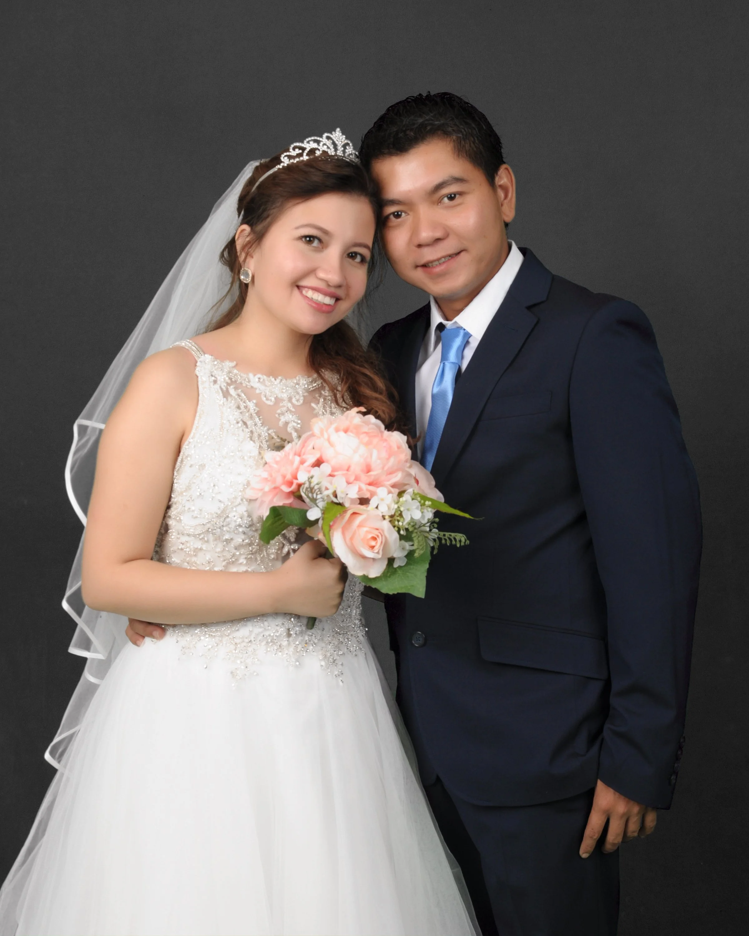 A bride and groom smiling, embracing, with the bride holding a bouquet of pink and white flowers, against a dark background.