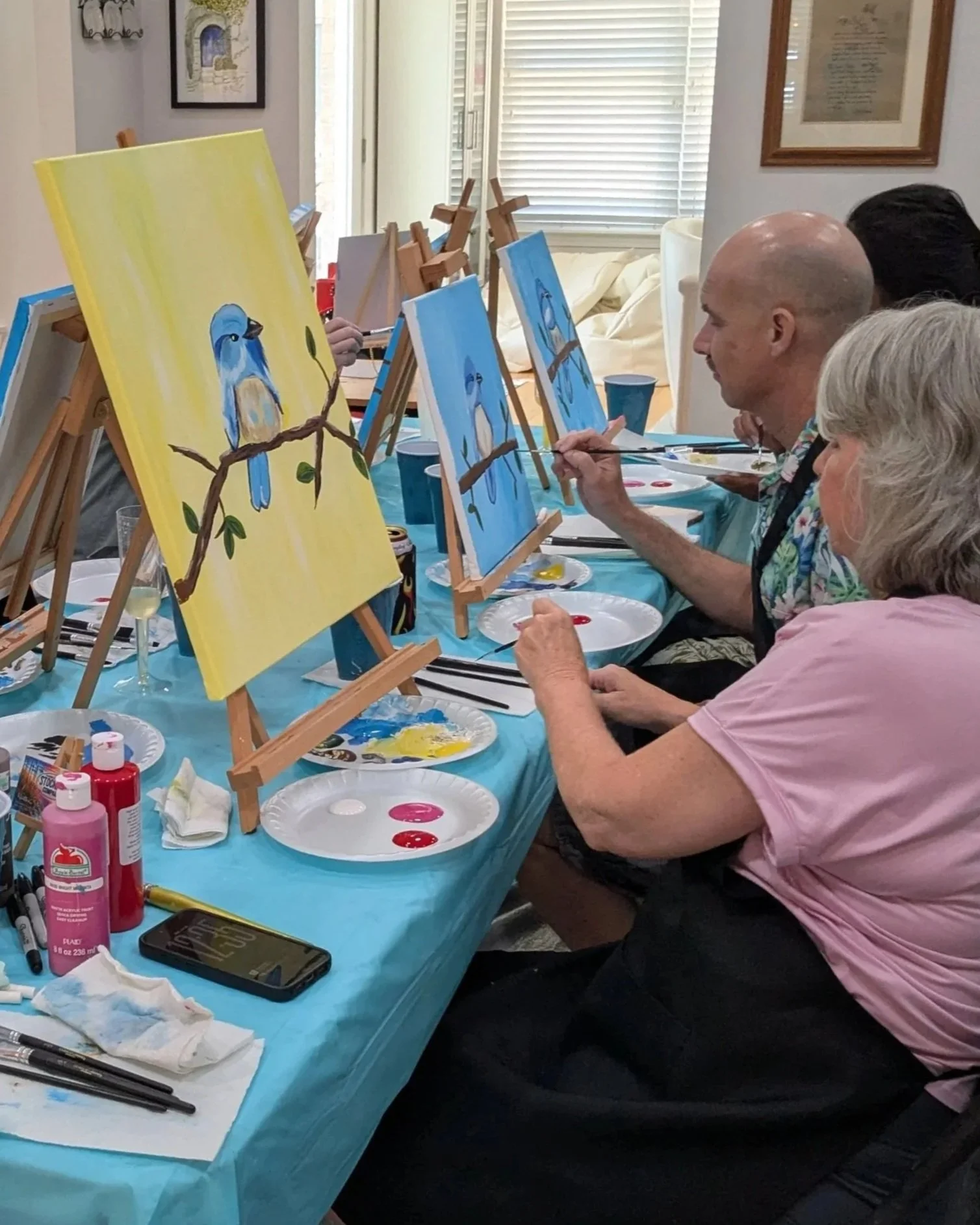 People painting blue and yellow bird on canvases at a painting class in a room with windows and framed art on the walls.