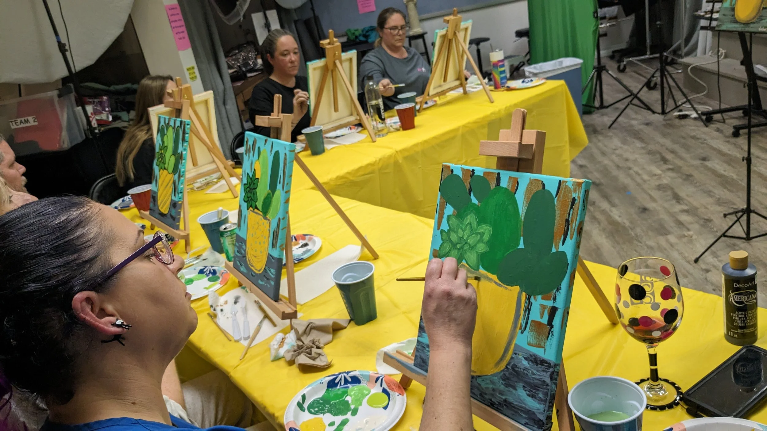Several women participating in a group painting class, sitting at a long yellow table with painted canvases and supplies. They are focused on their artwork, with easels holding colorful paintings of plants and fruits. The setting appears to be a studio or classroom, with art supplies, drinks, and equipment visible.