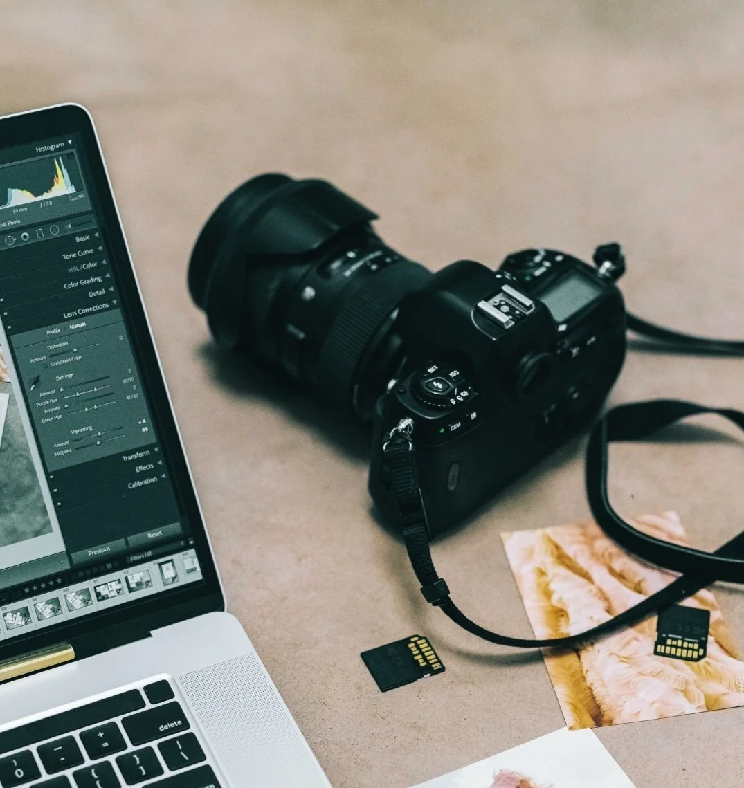 A digital camera connected to a laptop with photo editing software open, multiple SD cards, and printed photographs on a beige surface.