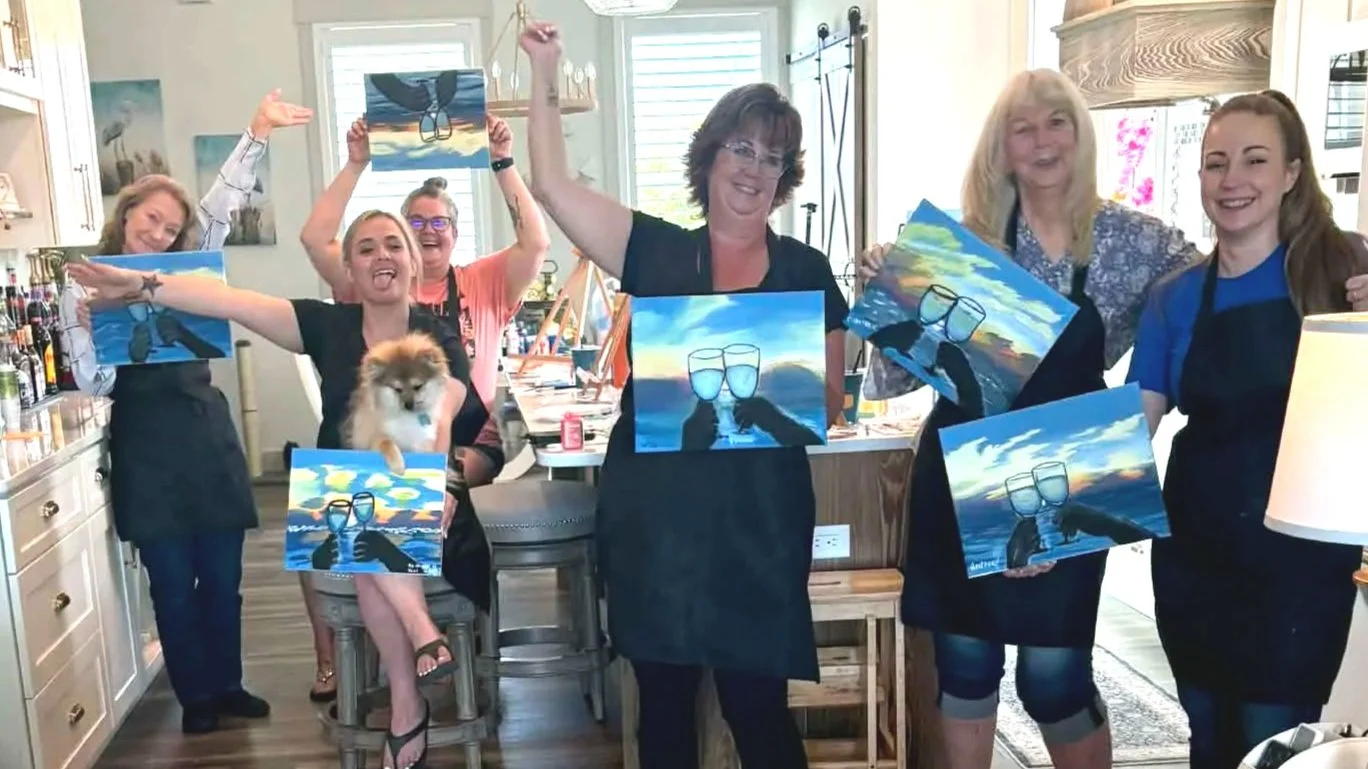 Group of women in a kitchen holding up paintings of glasses and a dog among a colorful sky and clouds, celebrating their art project.