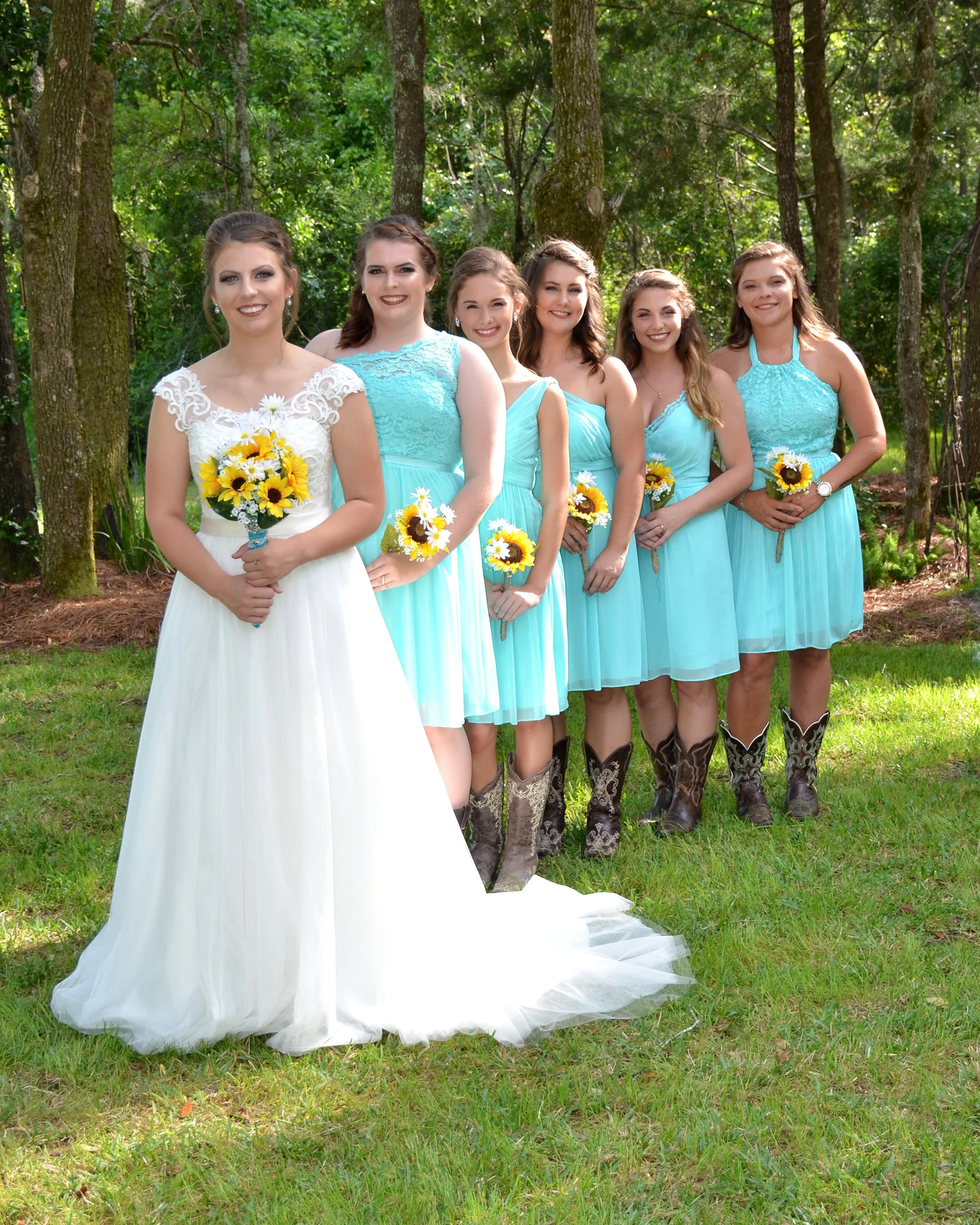 A bride and six bridesmaids standing outdoors in a wooded area, all holding sunflower bouquets. The bride is wearing a white wedding dress, and the bridesmaids are in matching light blue dresses with cowboy boots.