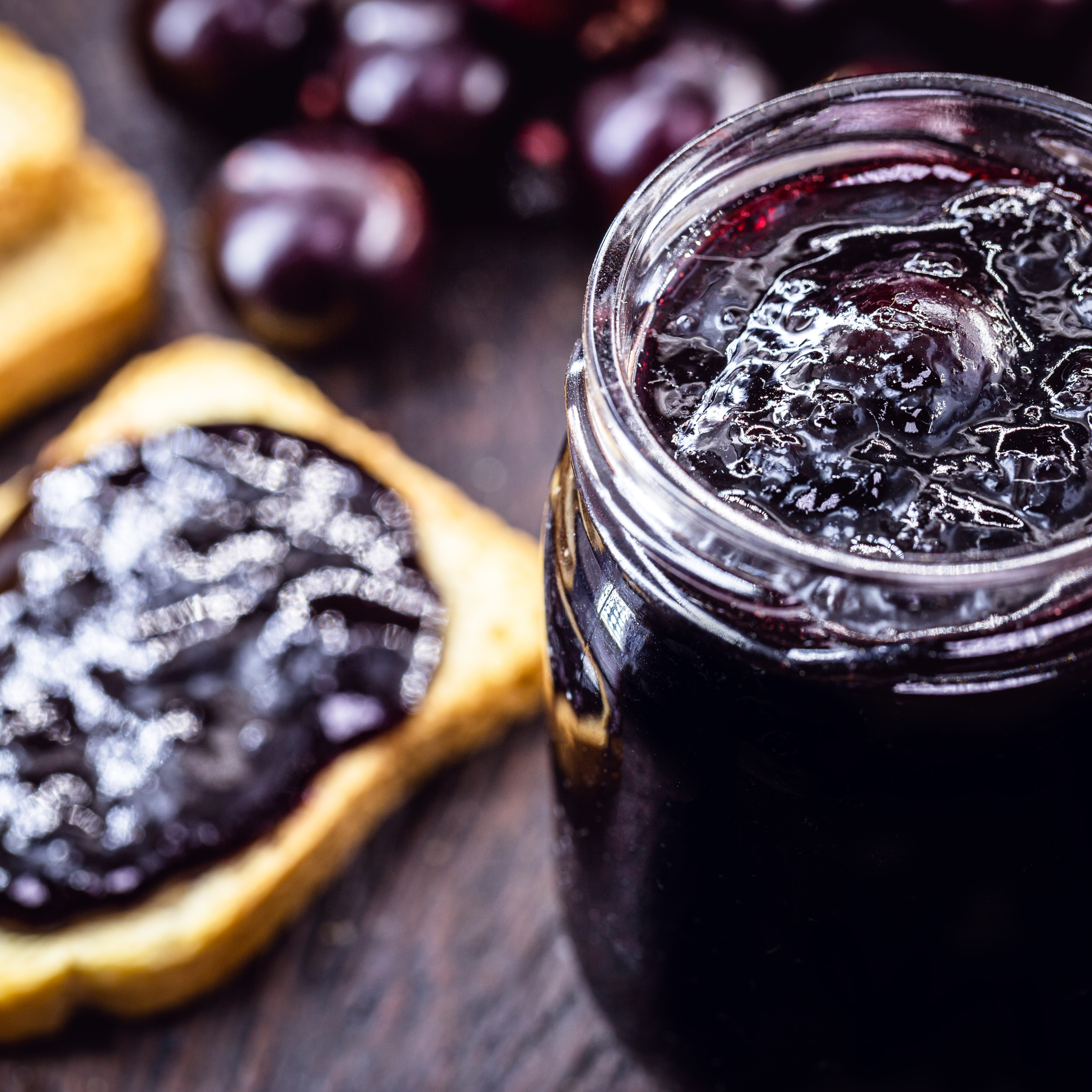 A glass jar filled with dark purple grape jam next to a slice of bread with grape jelly and some fresh grapes in the background.