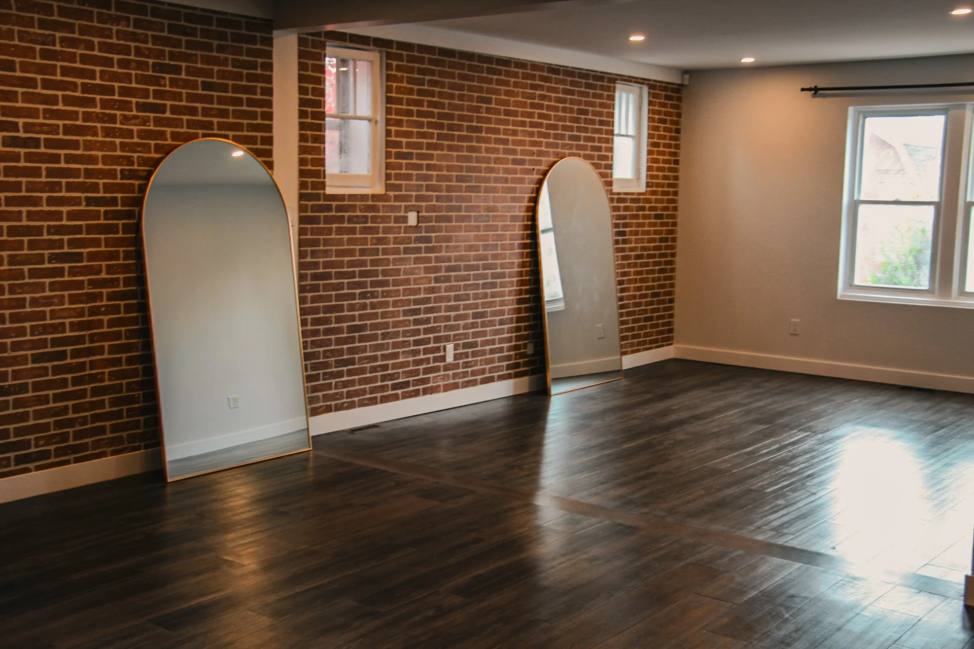 Empty room with dark wooden floors, brick wall with two small windows, beige wall with a window, and two standing mirrors leaning against the brick wall.