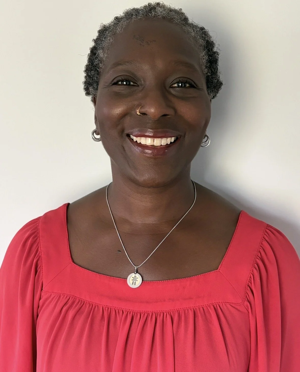 Smiling woman with short gray hair, wearing a red top, jewelry, and a necklace, standing against a white background.