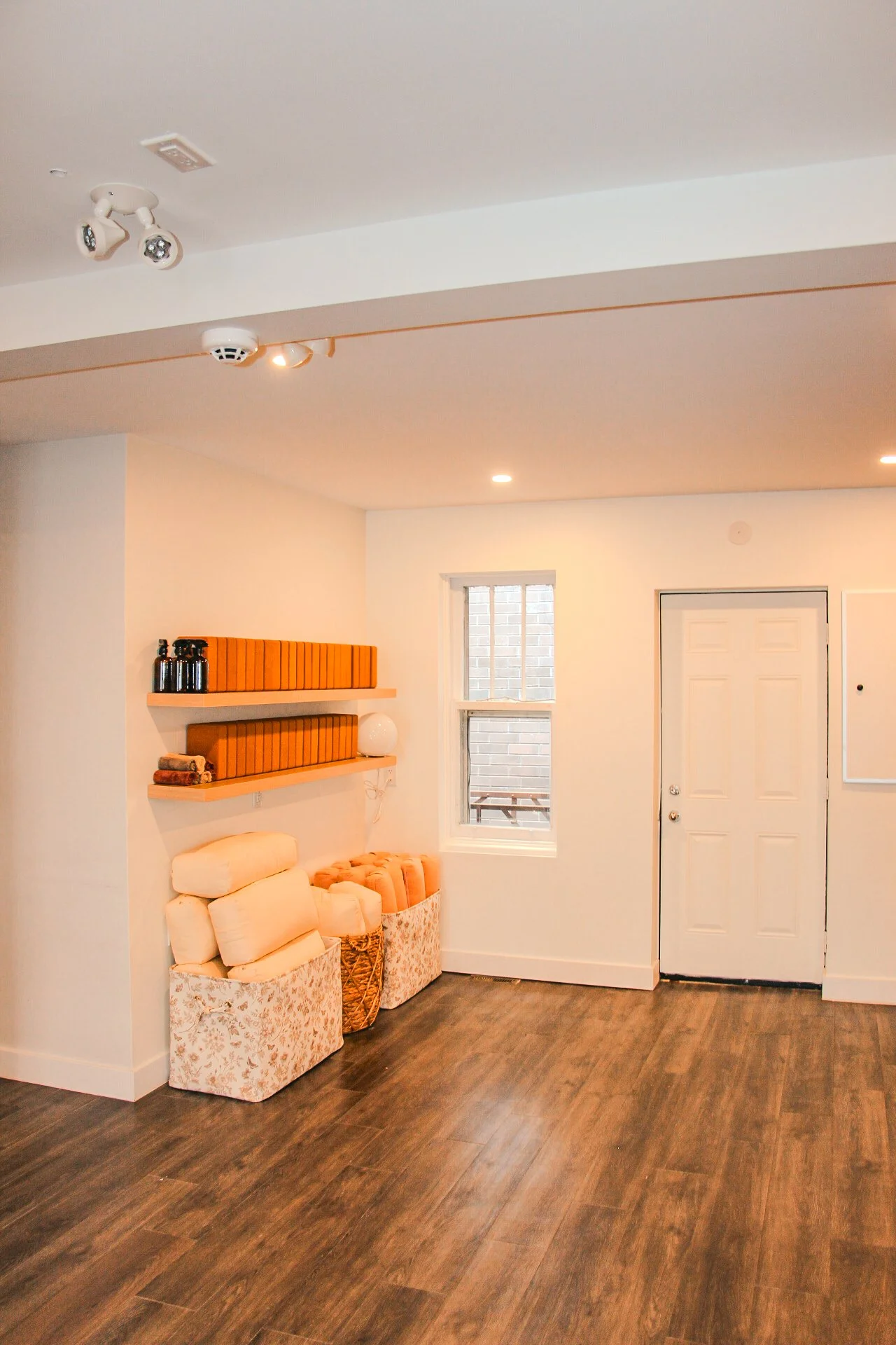 Living room with white walls, hardwood floor, window, white door, mounted shelves with orange books, decorative items, and folded blankets in storage baskets.