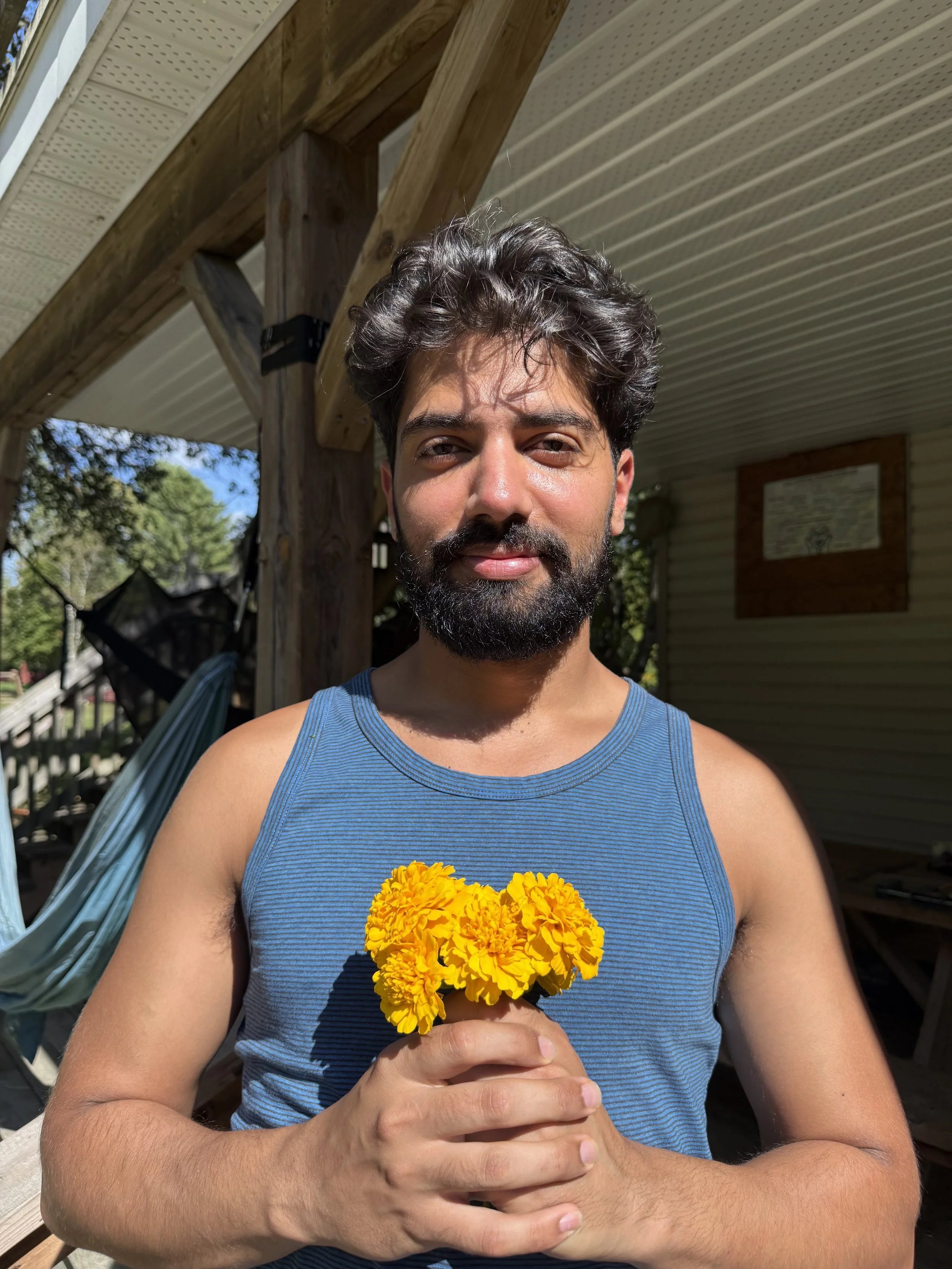 A man with dark, curly hair and a beard holding a small bouquet of yellow marigold flowers, standing outdoors in sunlight with a wooden structure and trees in the background.