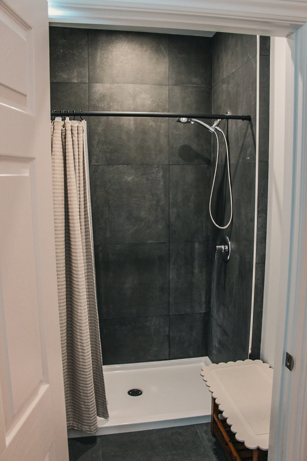 A modern shower with large dark gray tiles, a silver showerhead with a hose, a curtain on a black rod, and a white shower base. A small wooden tray with scalloped edge is placed outside the shower.