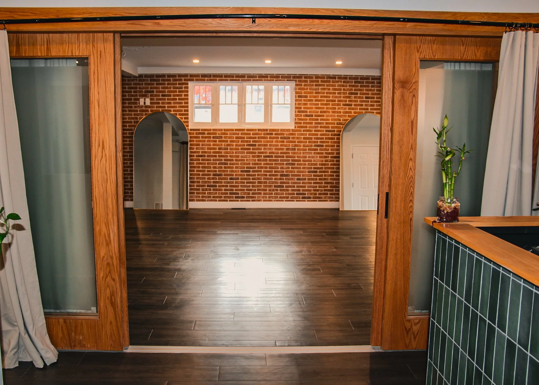 View of an interior room with exposed brick walls, three windows at the top, arched doorways, dark wooden flooring, and a potted plant on a tiled counter.