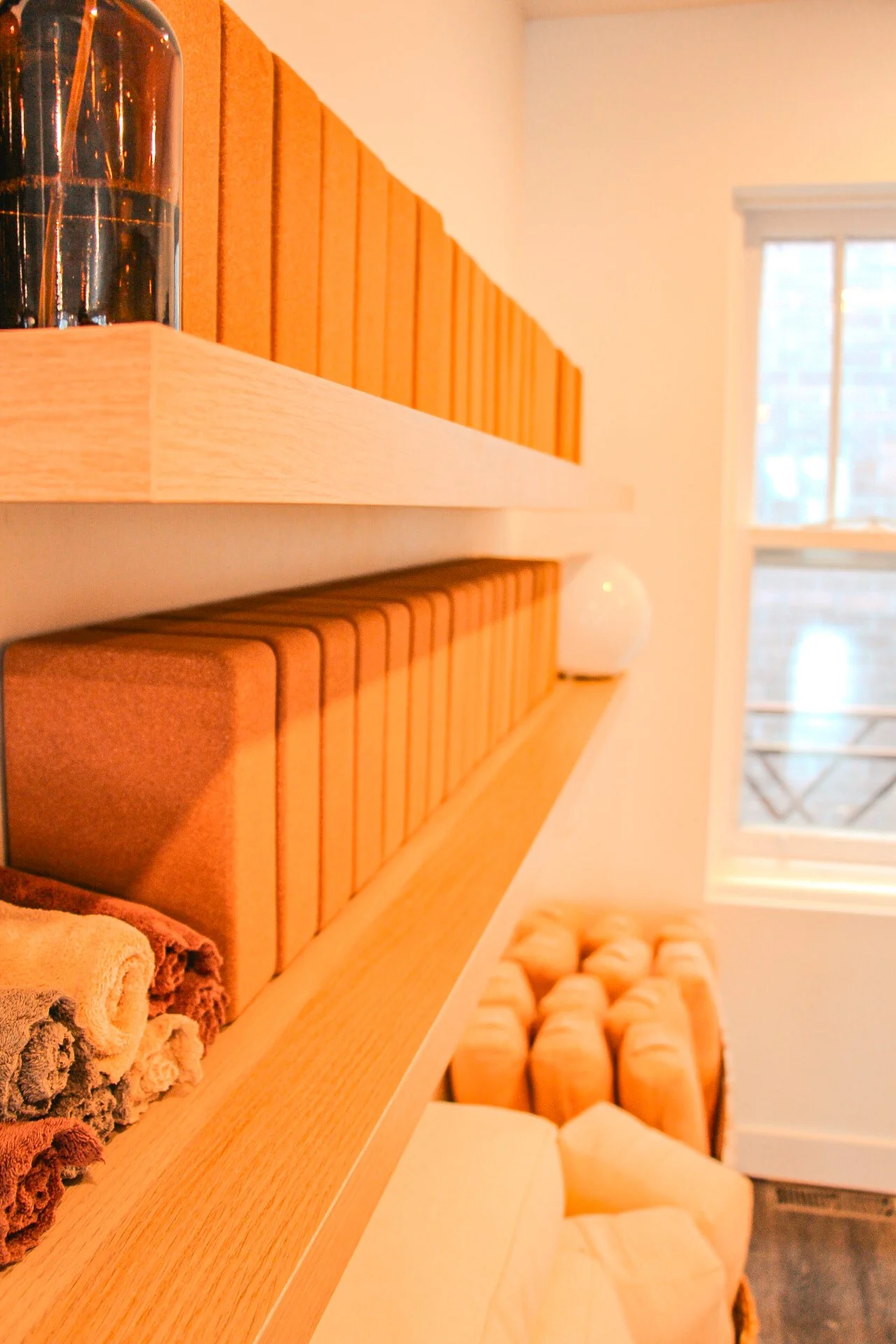 Close-up of a wooden shelf with orange books, towels, and a spherical object, next to a window with a view of a brick building outside.