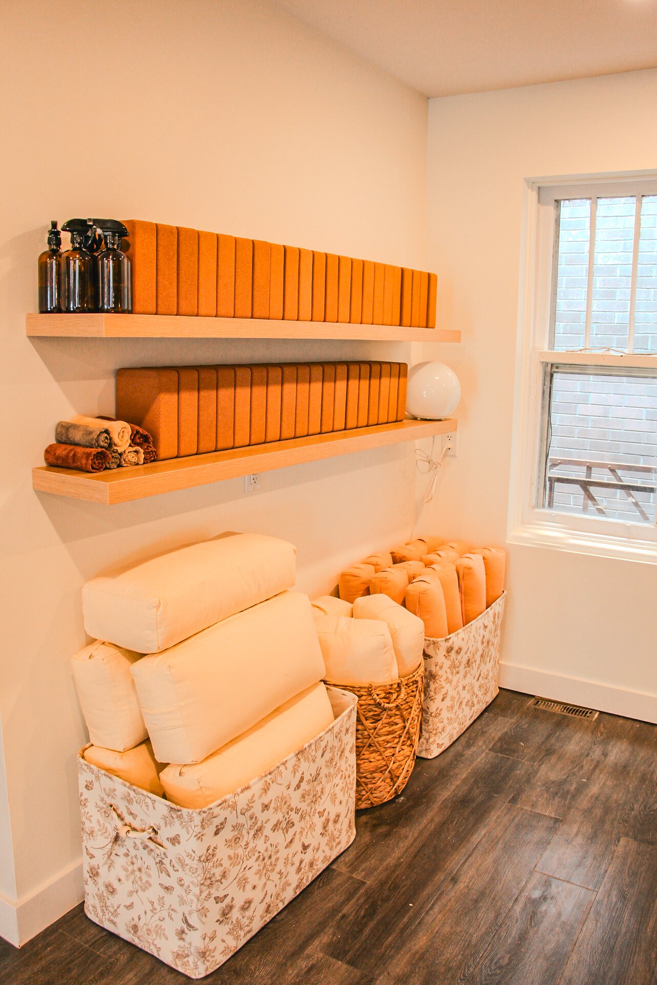 A cozy corner with two floating wooden shelves holding rolled towels, decorative boxes, spray bottles, and a lamp, next to a window and storage baskets filled with cushions and yoga bolsters on a dark wood floor.