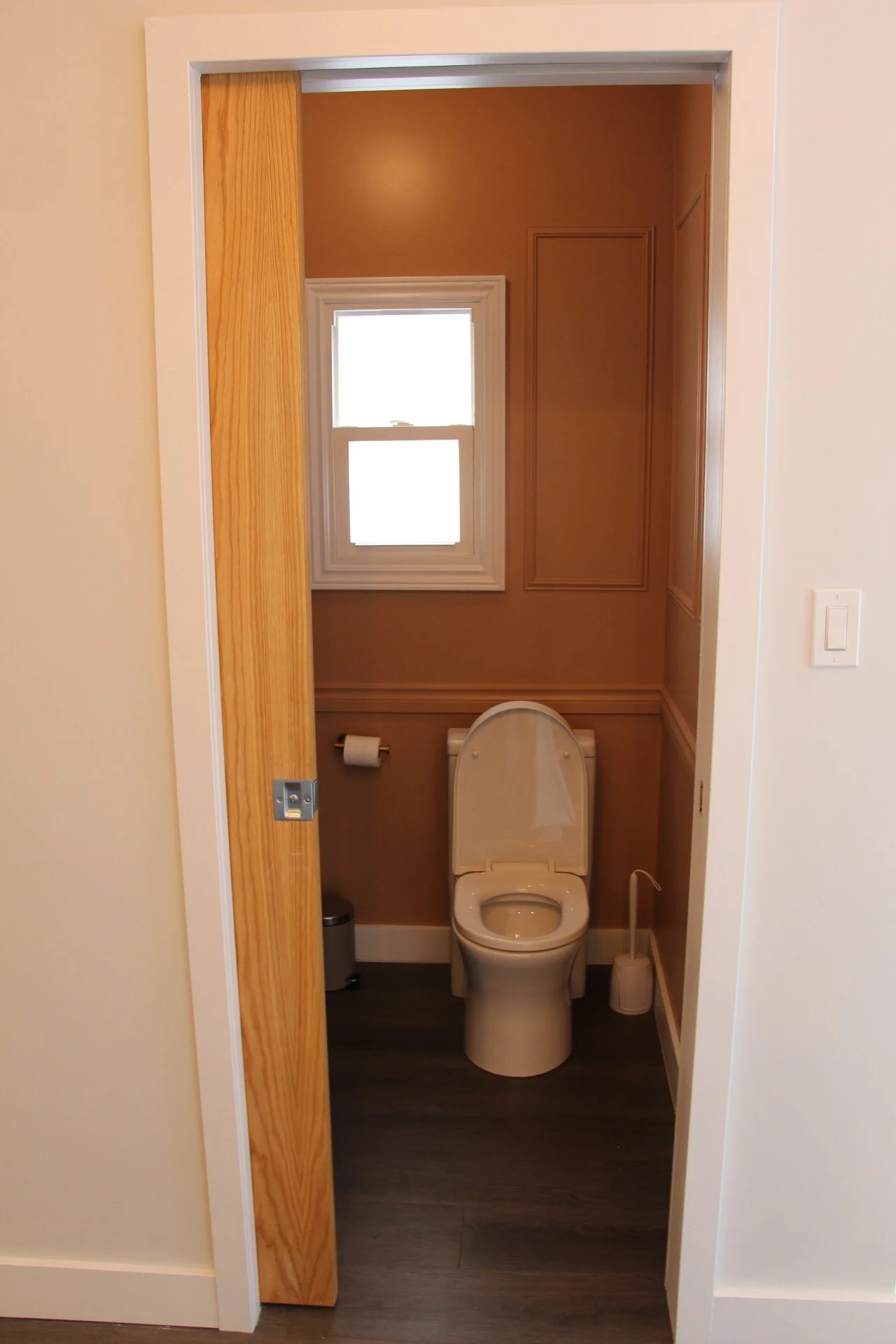 A small bathroom with toilet, window, toilet paper roll, trash can, and a toilet brush. The door is partially open, revealing a wooden door frame and beige walls with wallpaper. There is a framed paneled design on the wall and dark hardwood flooring.