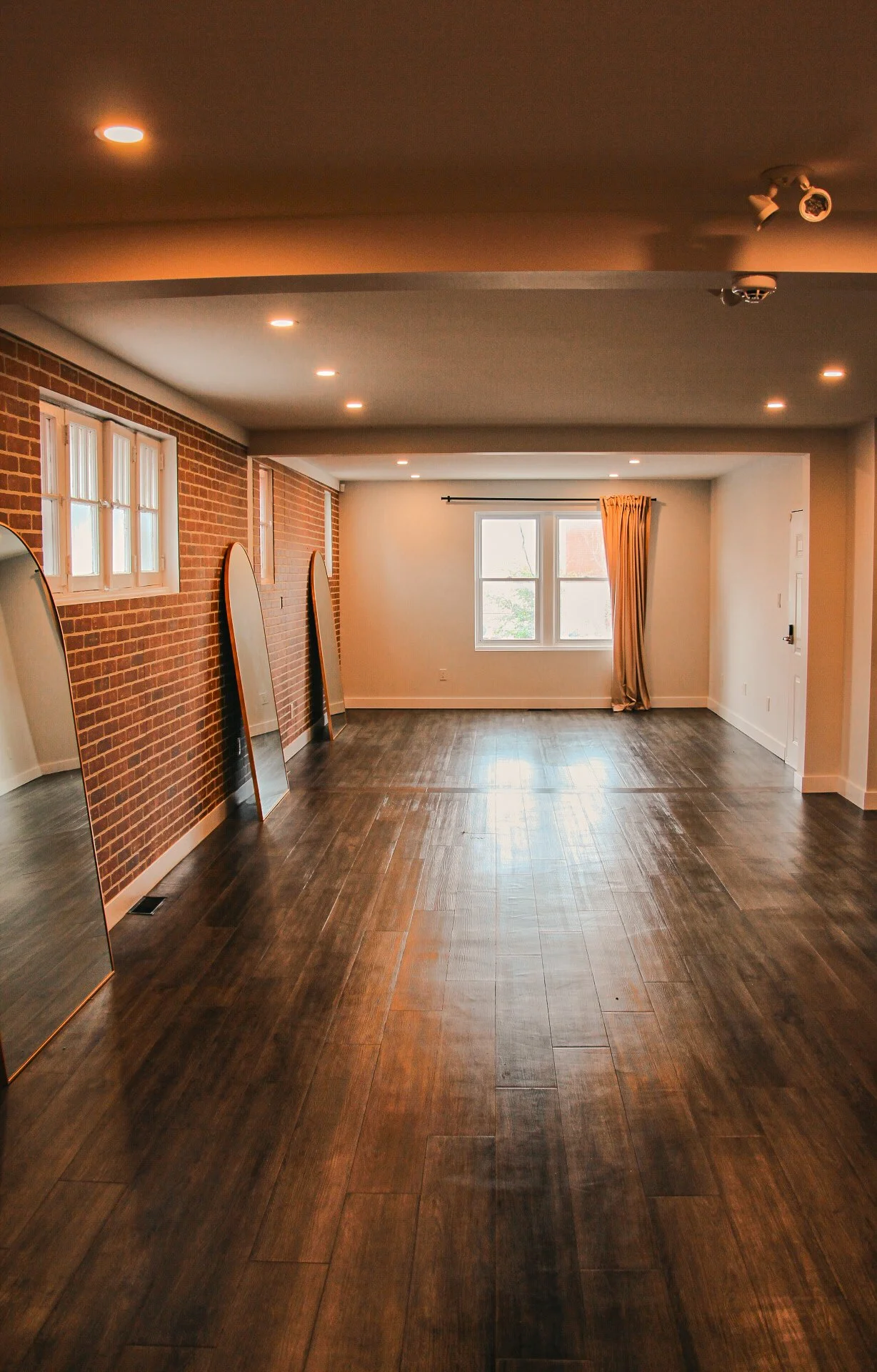 Empty room with dark wood flooring, brick accent wall with windows, and mirrors leaning against the wall.