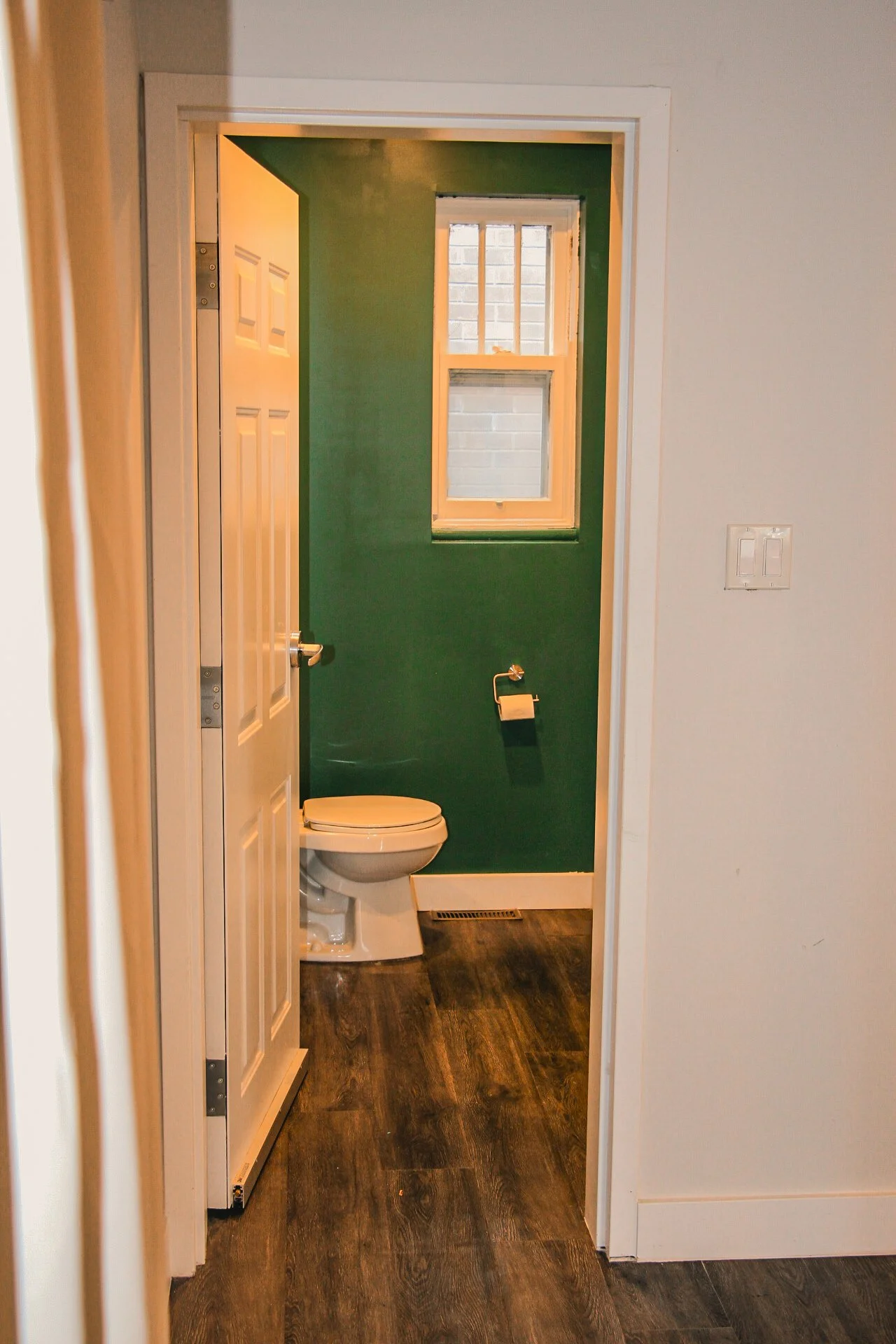 Small bathroom with a green wall, a window, a toilet, a toilet paper holder, and dark wood flooring.
