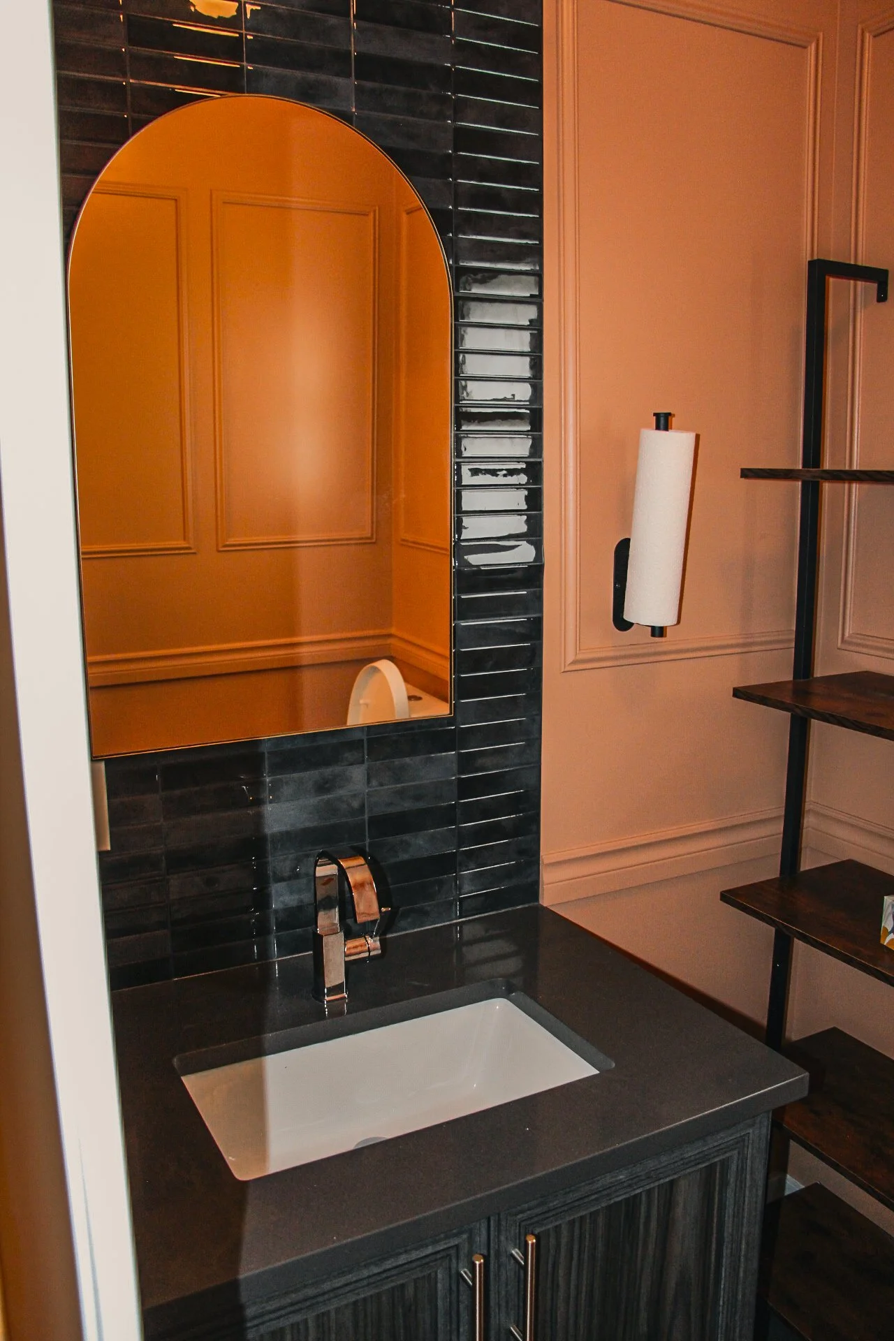 A modern bathroom vanity with a rectangular white sink, black countertop, black tile backsplash, a tall oval mirror, and a paper towel holder mounted on the wall. Wooden shelves are visible on the right.