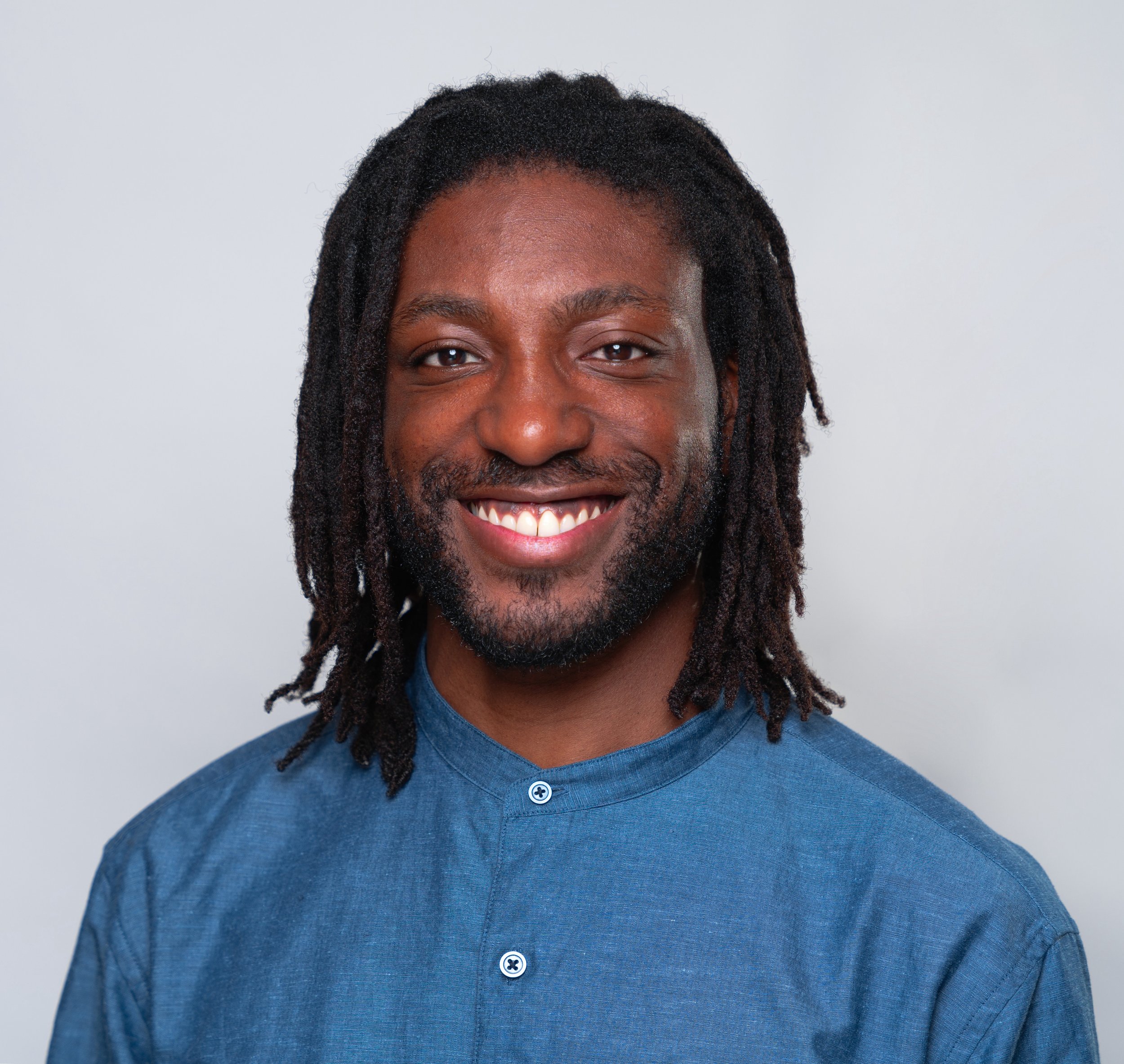 A smiling man with dreadlocks, a beard, and a blue collared shirt against a plain light background.