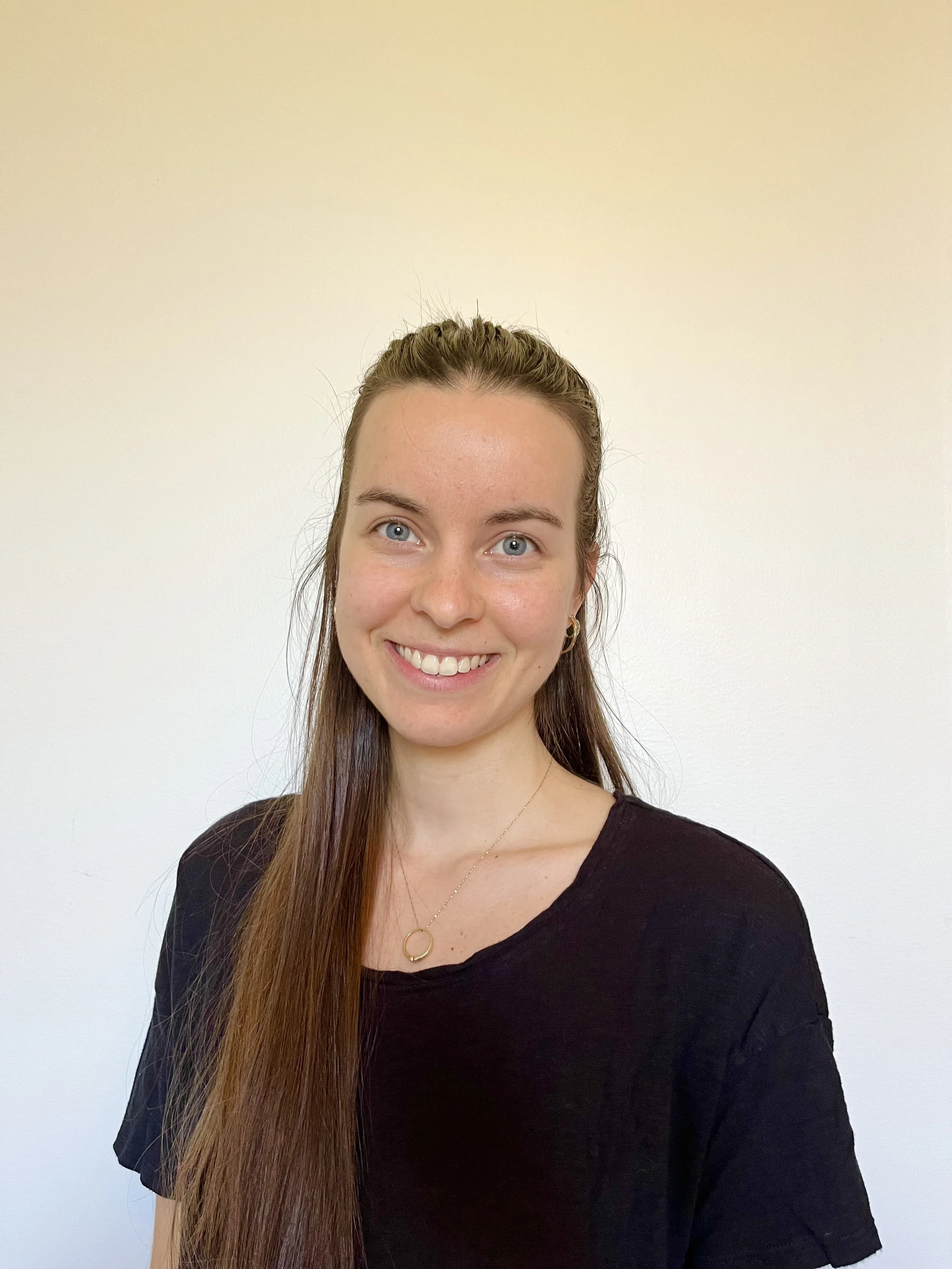 A young woman with long brown hair, blue eyes, and fair skin smiling, wearing a black t-shirt and gold jewelry, standing against a plain white background.