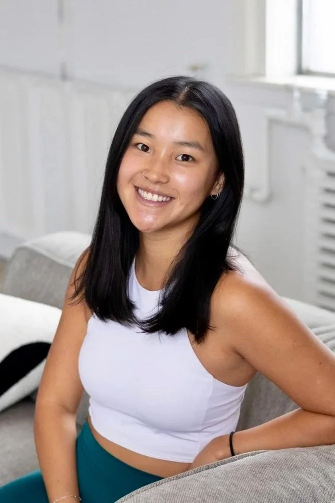 Smiling young woman with long black hair wearing a white sleeveless crop top and teal pants, sitting on a beige sofa in a bright room.