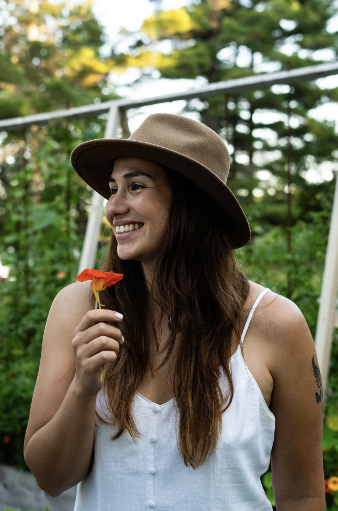 A smiling woman with long brown hair wearing a tan wide-brimmed hat and a white sleeveless top, holding a bright orange flower in her hand outdoors with green trees in the background.