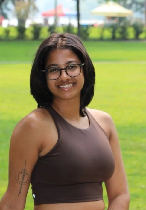 A woman with shoulder-length dark hair, glasses, and a tan tank top smiling outdoors on a grassy field