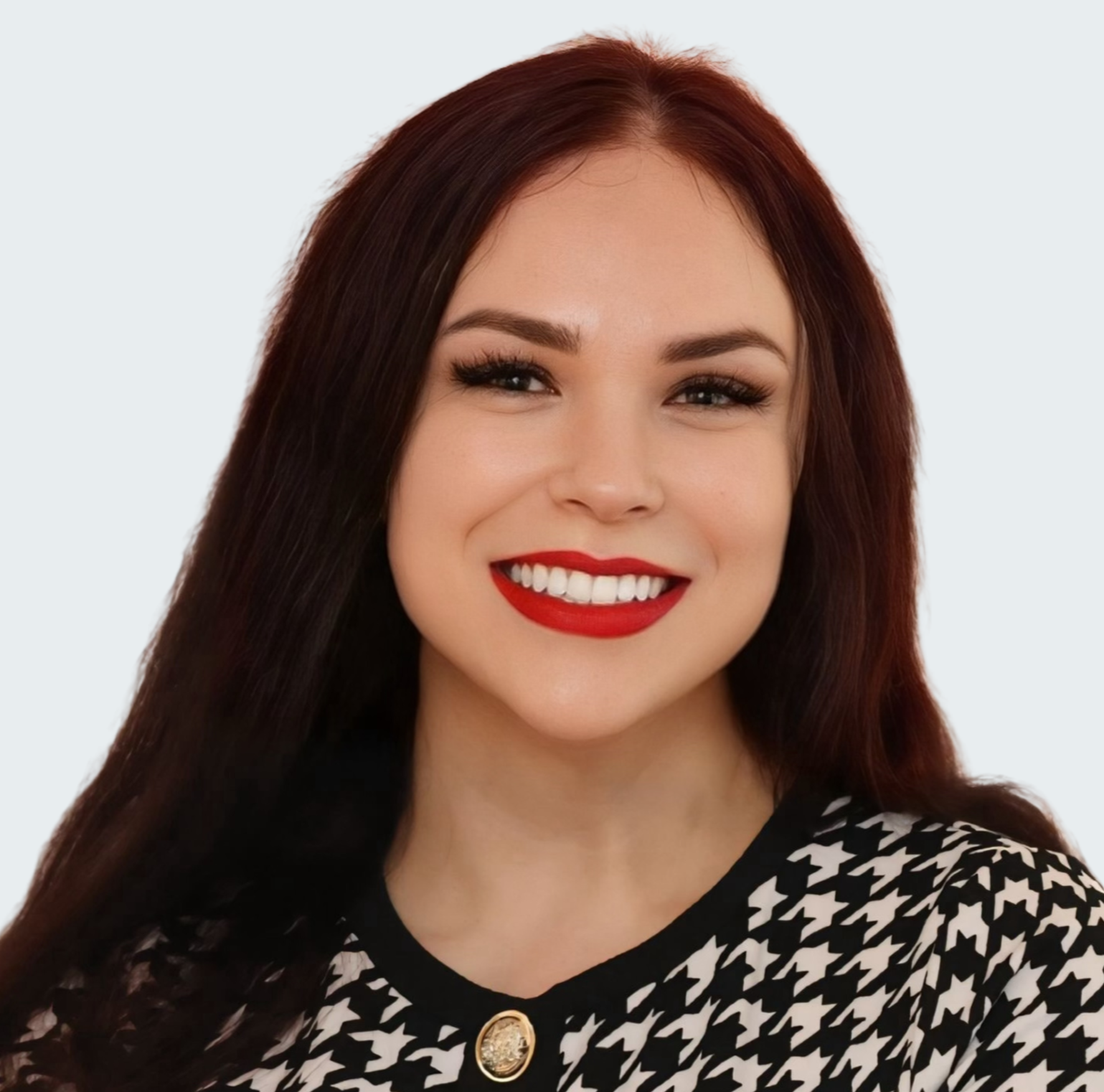 A woman with long dark purple hair, wearing red lipstick, smiling, and dressed in a blue sweater against a plain light background.