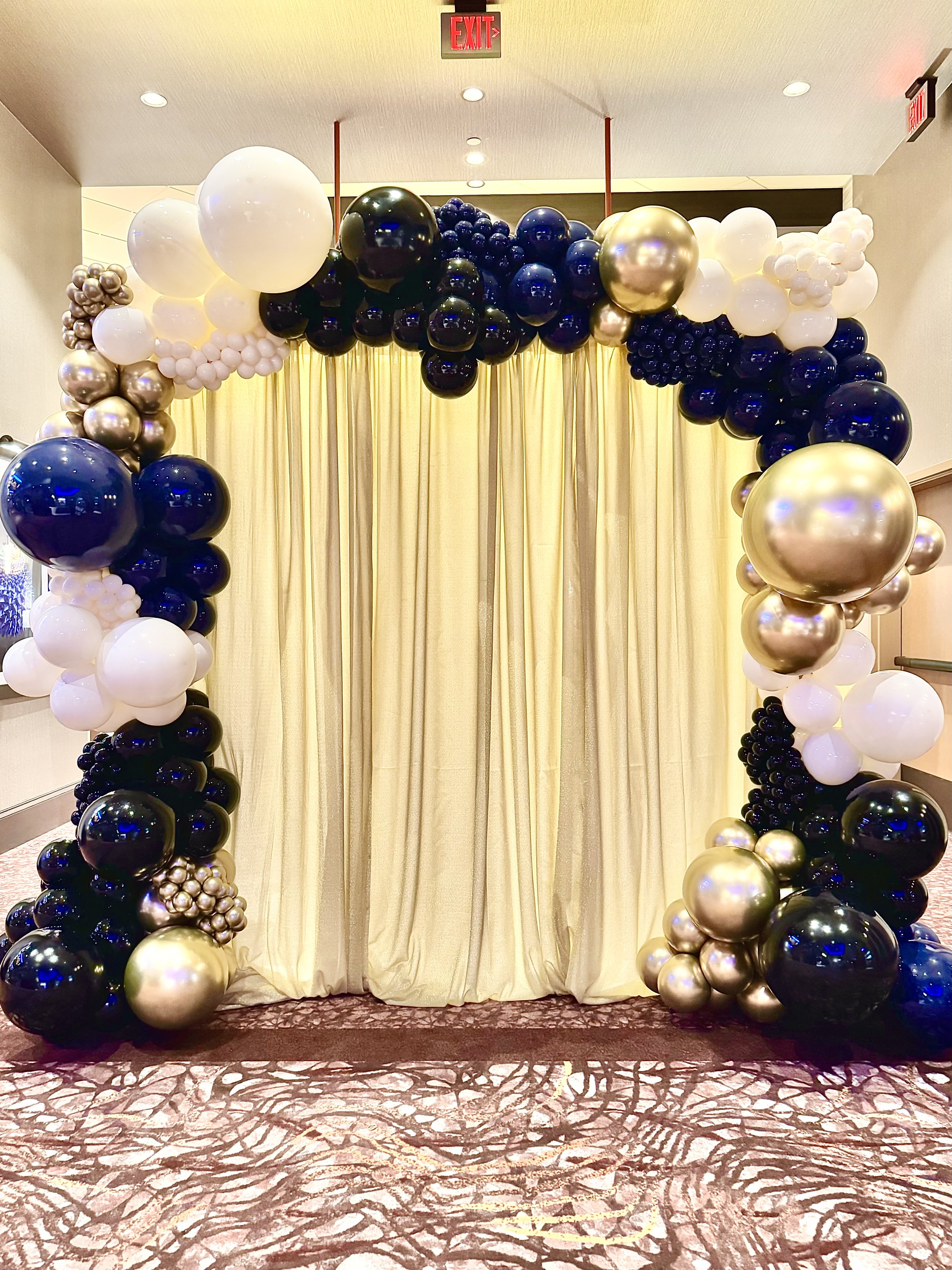 Balloon arch with white, black, gold, and metallic balloons framing a gold curtain backdrop.