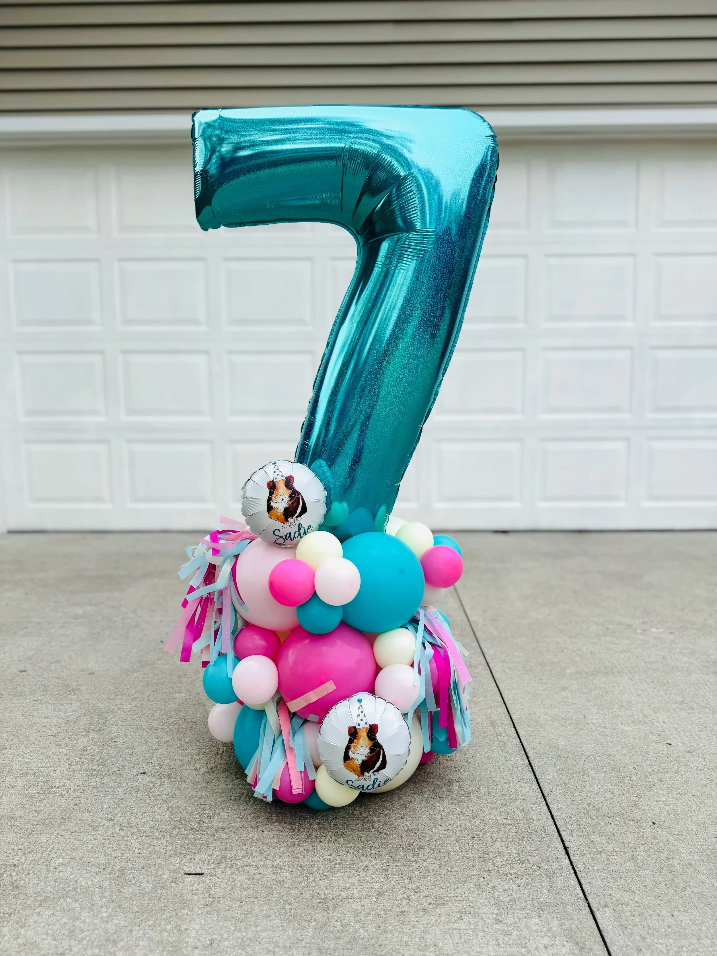 Large blue number 7 balloon surrounded by pink, blue, white, and cream smaller balloons with two small foil balloons featuring a guinea pig and the name Sadie, and colorful paper tassels at the base, on a concrete driveway in front of a garage.