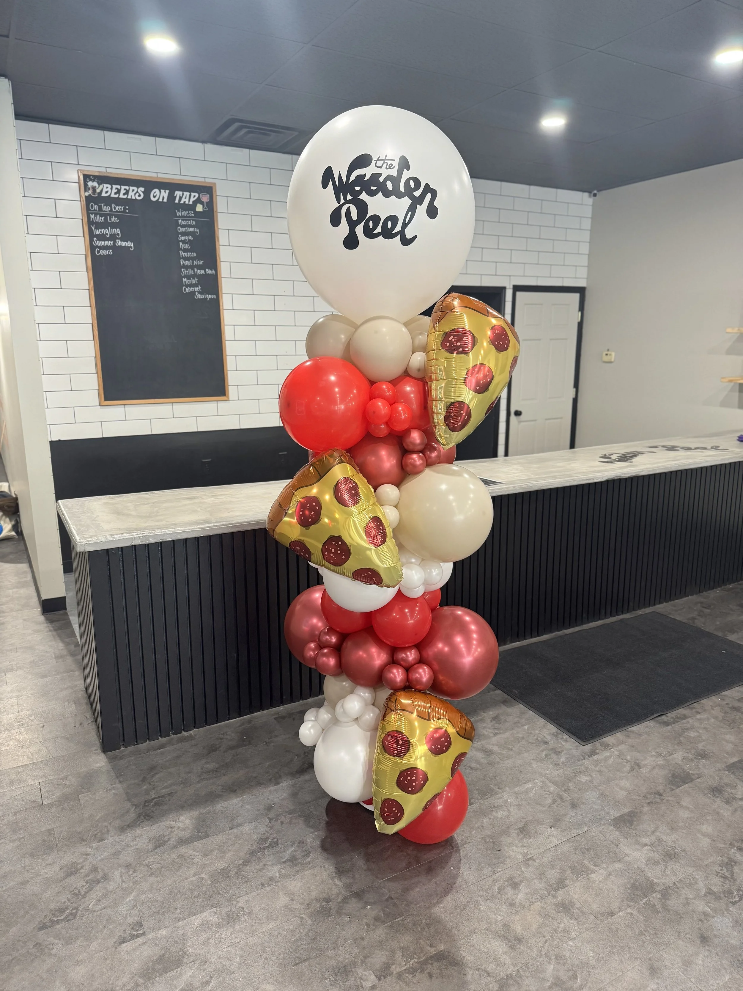 Balloon arrangement featuring large white balloons with the logo 'The Noodle Peel', smaller white and red balloons, and pizza slice-shaped balloons with pepperoni toppings, set against a restaurant interior background.