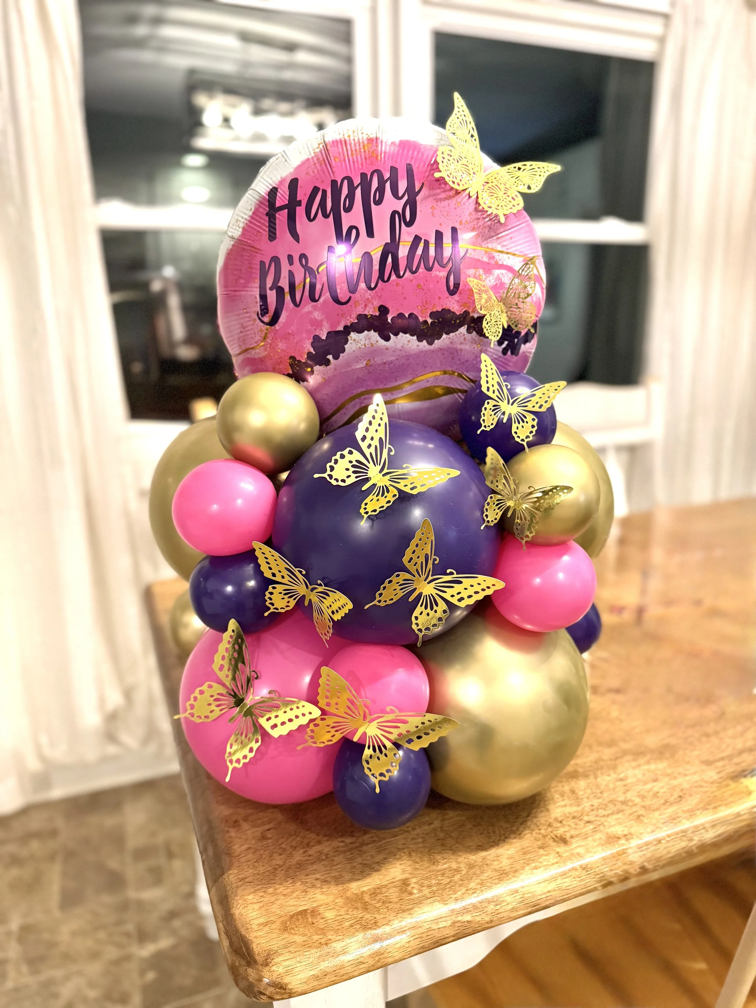 Colorful balloon bouquet with pink, purple, and gold balloons, decorated with gold butterfly confetti and a pink birthday sign reading 'Happy Birthday.'