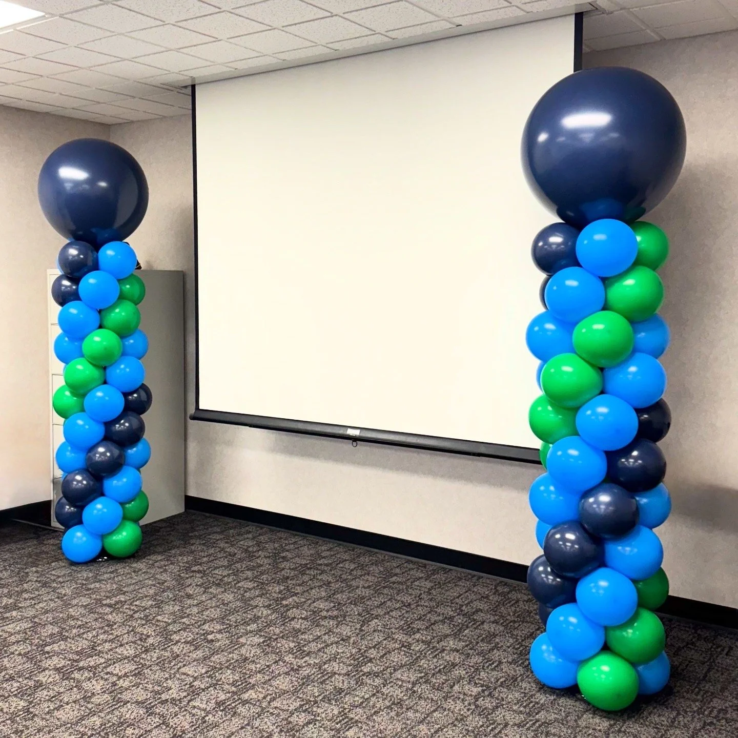 Decorative balloon columns with large dark blue balloons on top and smaller blue, green, and dark blue balloons below, in front of a white projection screen in a conference room.