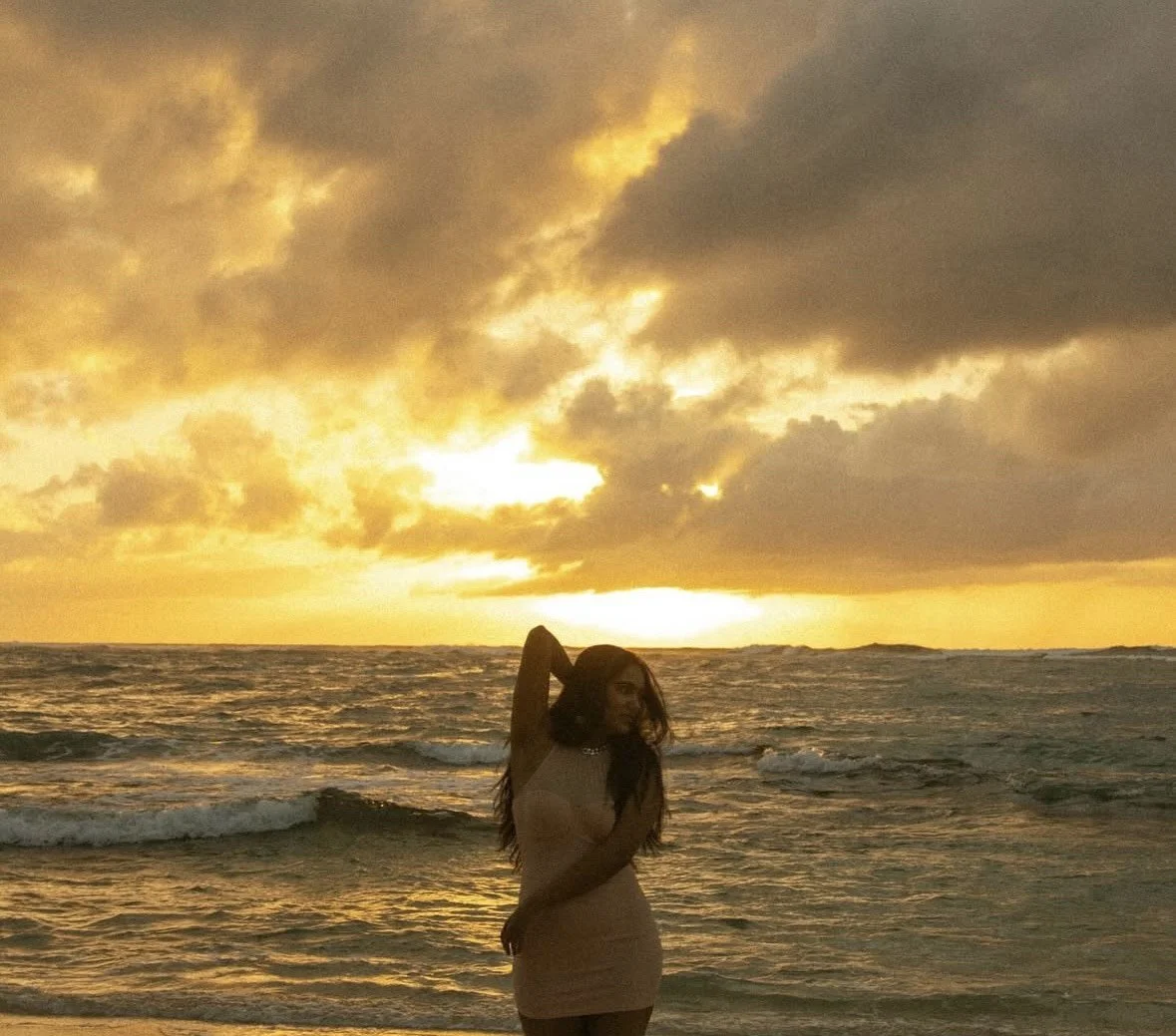 A woman standing on the beach during sunset with dark clouds overhead and the ocean behind her.