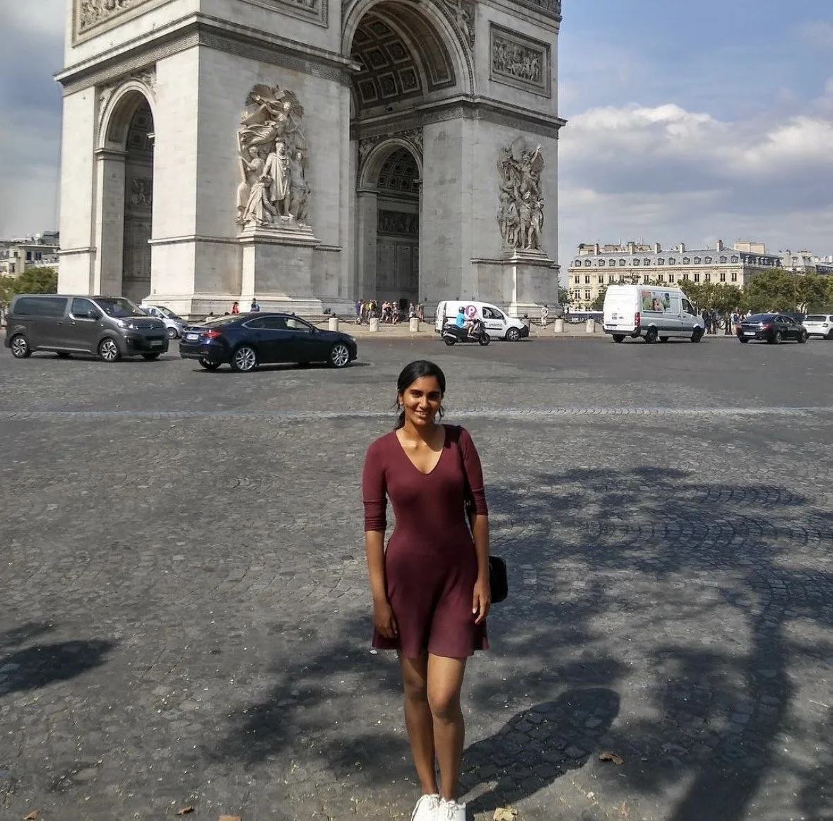 A woman stands on a cobblestone street in front of the Arc de Triomphe in Paris, France, with cars and motorcycles in the background under a partly cloudy sky.