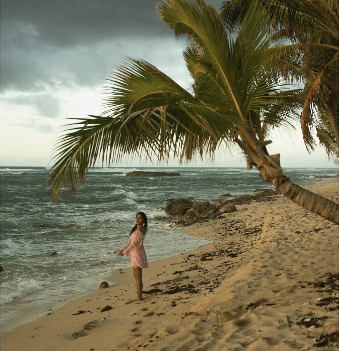 A woman in a pink dress standing on a sandy beach under a palm tree, with the ocean and rocks in the background on a cloudy sky day.
