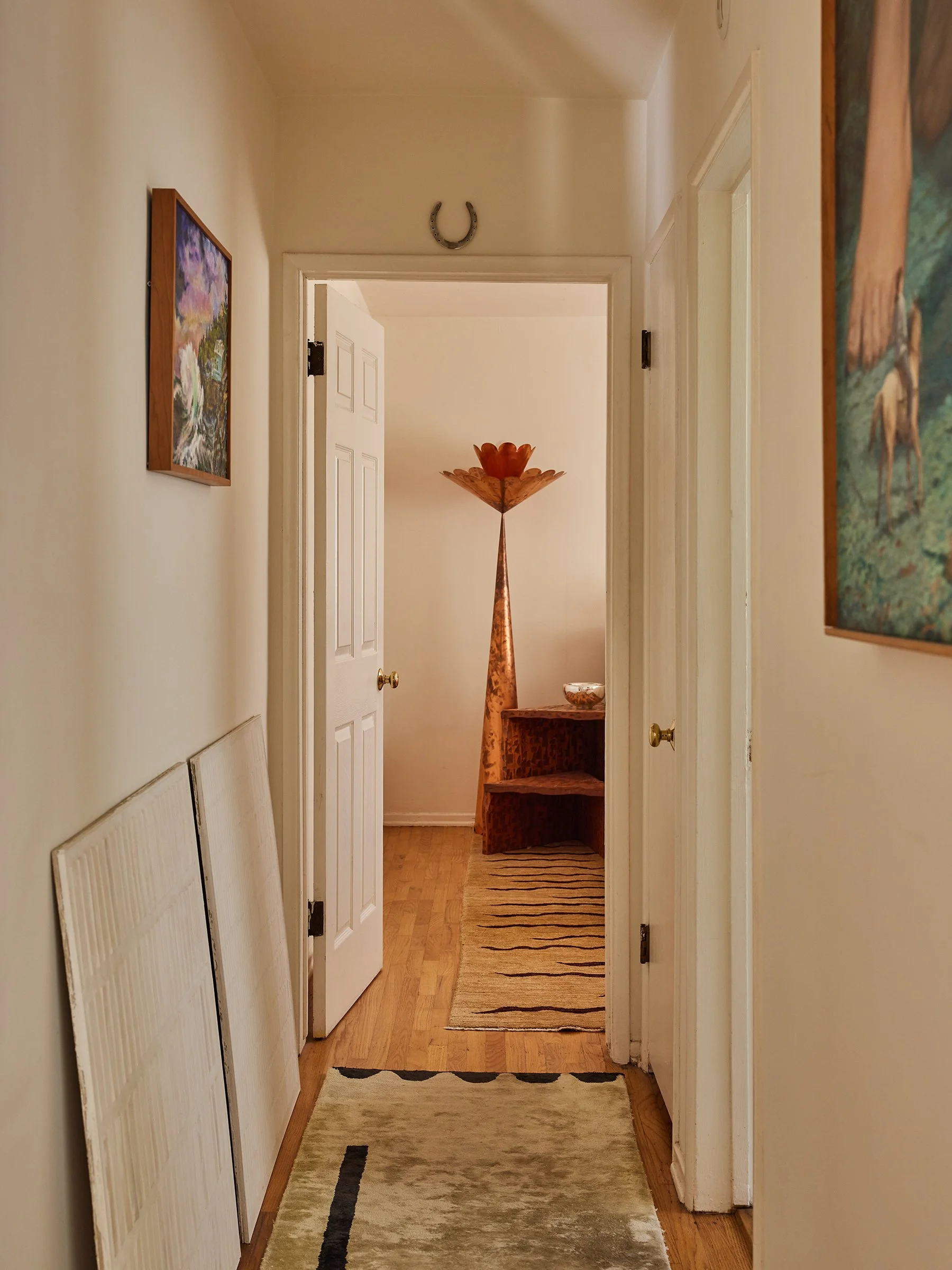 A hallway with a horseshoe near the ceiling, two framed paintings, a folding screen leaning against the wall on the left, a rug on the wooden floor, and a tall decorative lamp with a flower-shaped top in the background.
