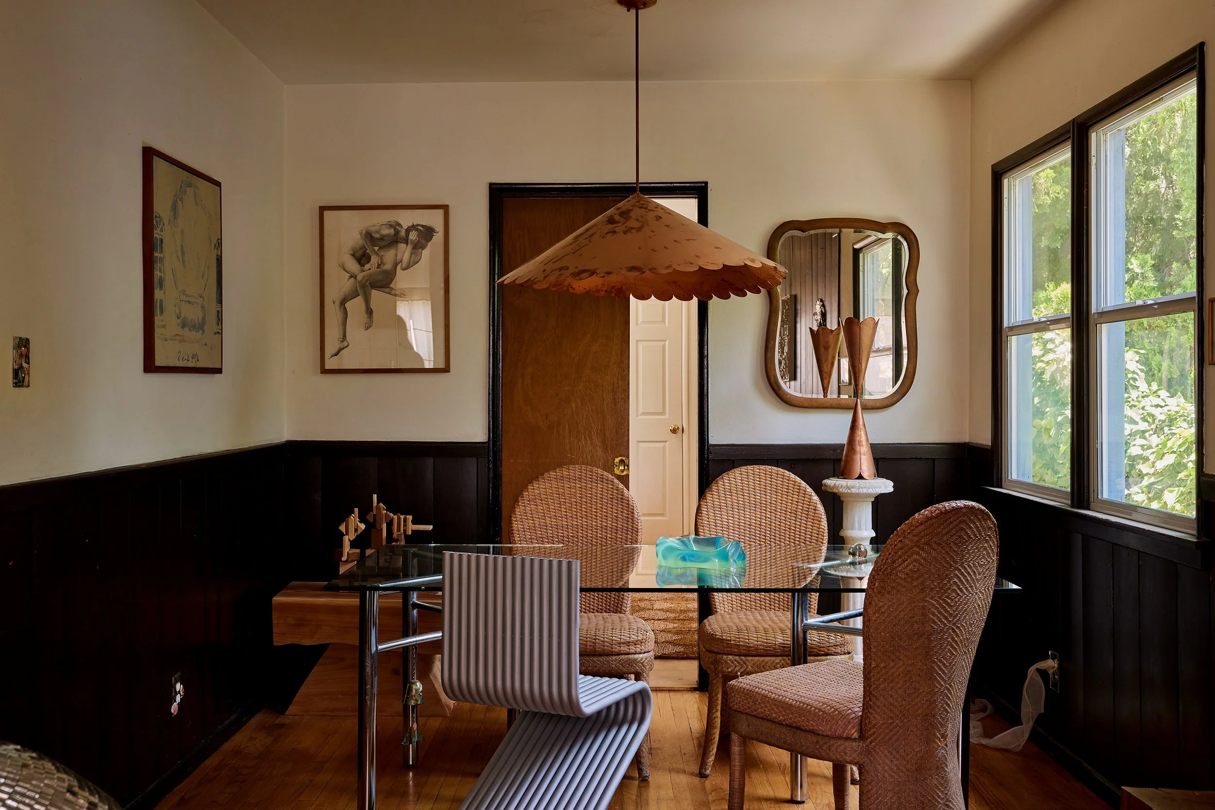 Vintage dining room with a glass table, mixed chairs, wall art, a mirror, and large windows showing green trees outside.