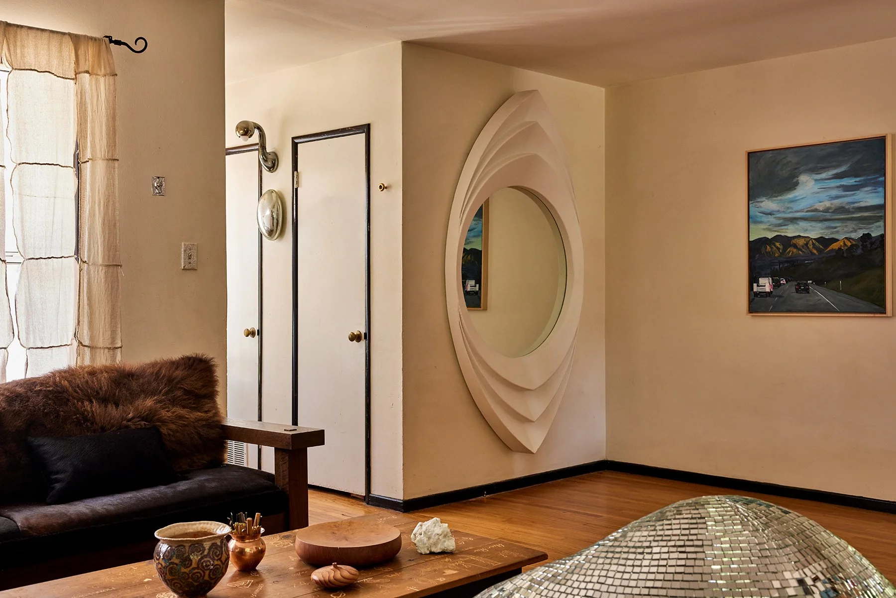 Living room with a wooden coffee table, a black couch with a furry throw pillow, and a window with beige curtains. A circular mirror with a layered frame is on the wall, with a mountain landscape painting on the adjacent wall. There are two closed do