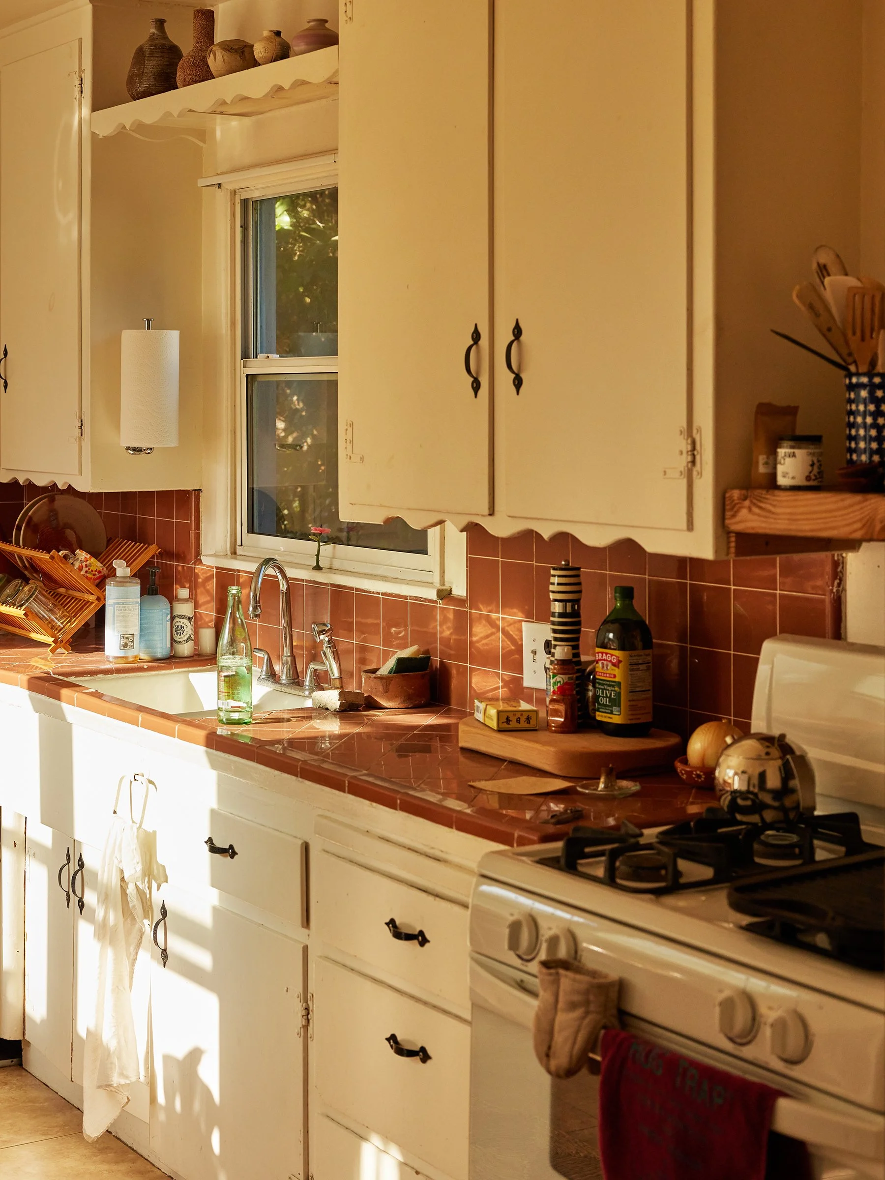 A cozy kitchen with white cabinets, a red tiled countertop, and a window letting in sunlight. There are kitchen utensils, bottles, and a stove with a towel hanging from it.