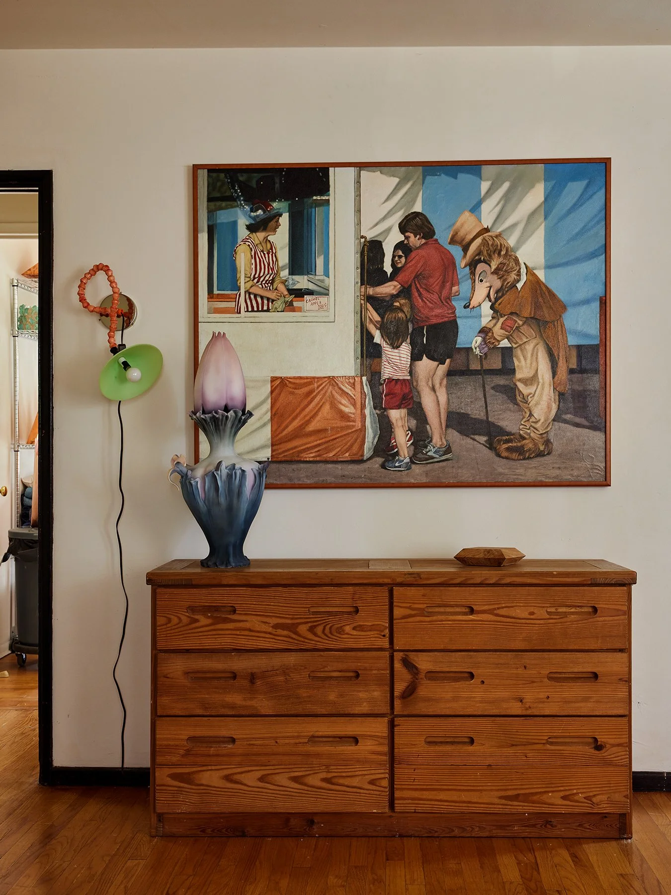 A wooden sideboard with a decorative vase and a small wooden dish on top, a colorful wall painting of children and a mascot at a carnival, and a wall-mounted lamp with a green shade and orange cord.
