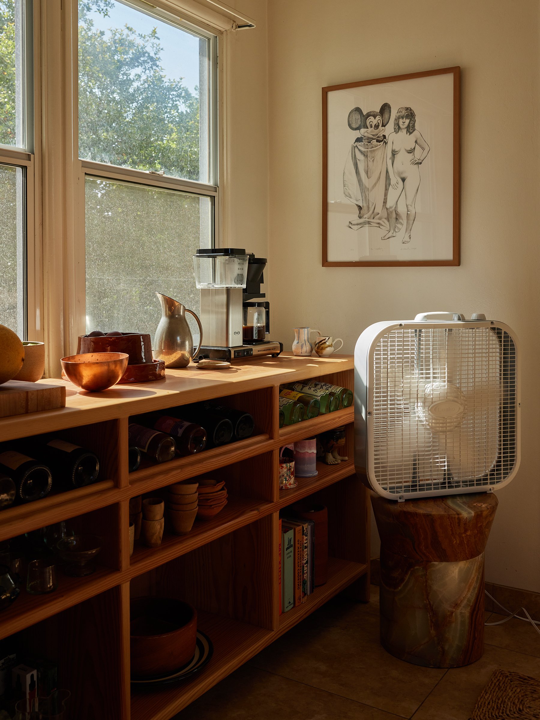 A corner of a room with a wooden shelf holding glassware, books, bowls, and a coffee machine, next to a window with trees outside. A framed drawing of a woman and a cartoon mouse hangs on the wall, and a large white fan rests on a marble-colored pede