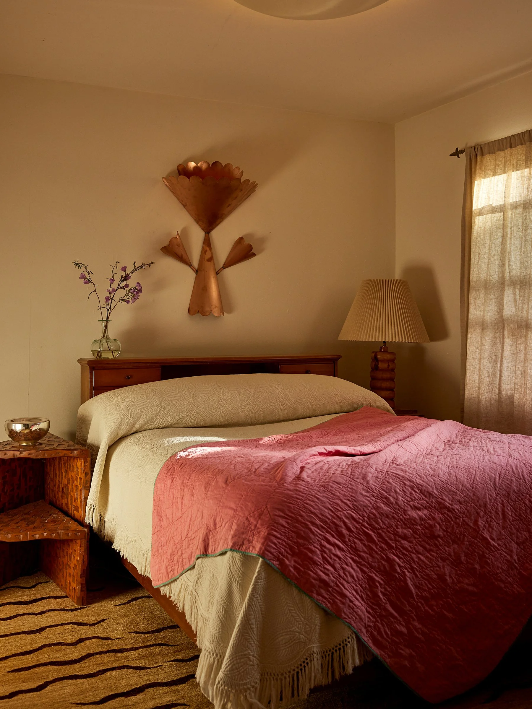 A cozy bedroom with a bed covered in a pink quilt and a cream-colored blanket, a wooden nightstand with a glass bowl, a table lamp, a window with beige curtains, a wall art piece, and a vase with purple flowers.