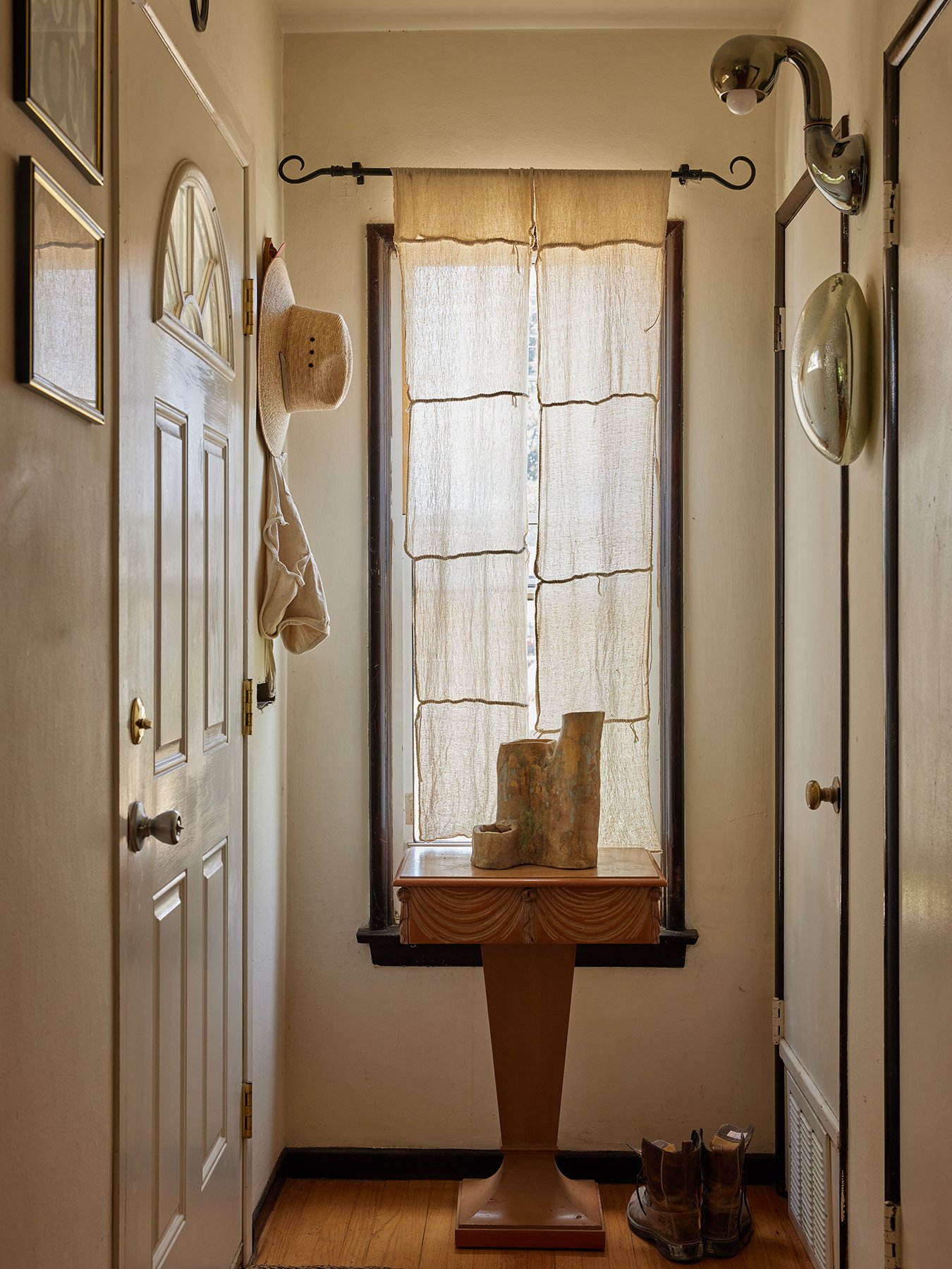 A narrow entryway with a window covered by beige curtains, a wooden table with a stone sculpture, a pair of brown boots on the floor, and hats and bags hanging on the wall.